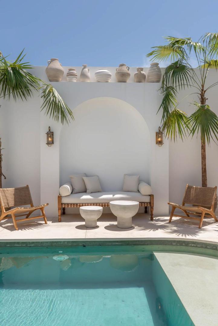 Built-in banquette and woven chairs under an arch at the head of the pool, palms behind