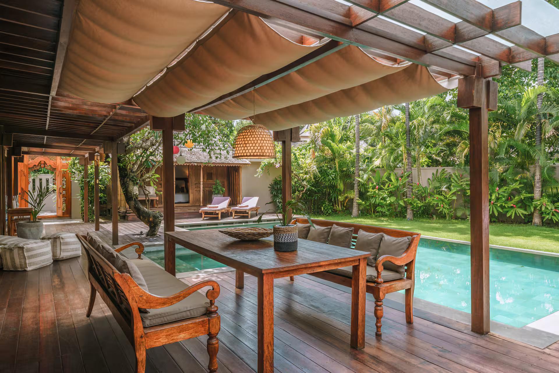 Teak dining table and carved bench under the canvas-shaded pergola at the pool's edge