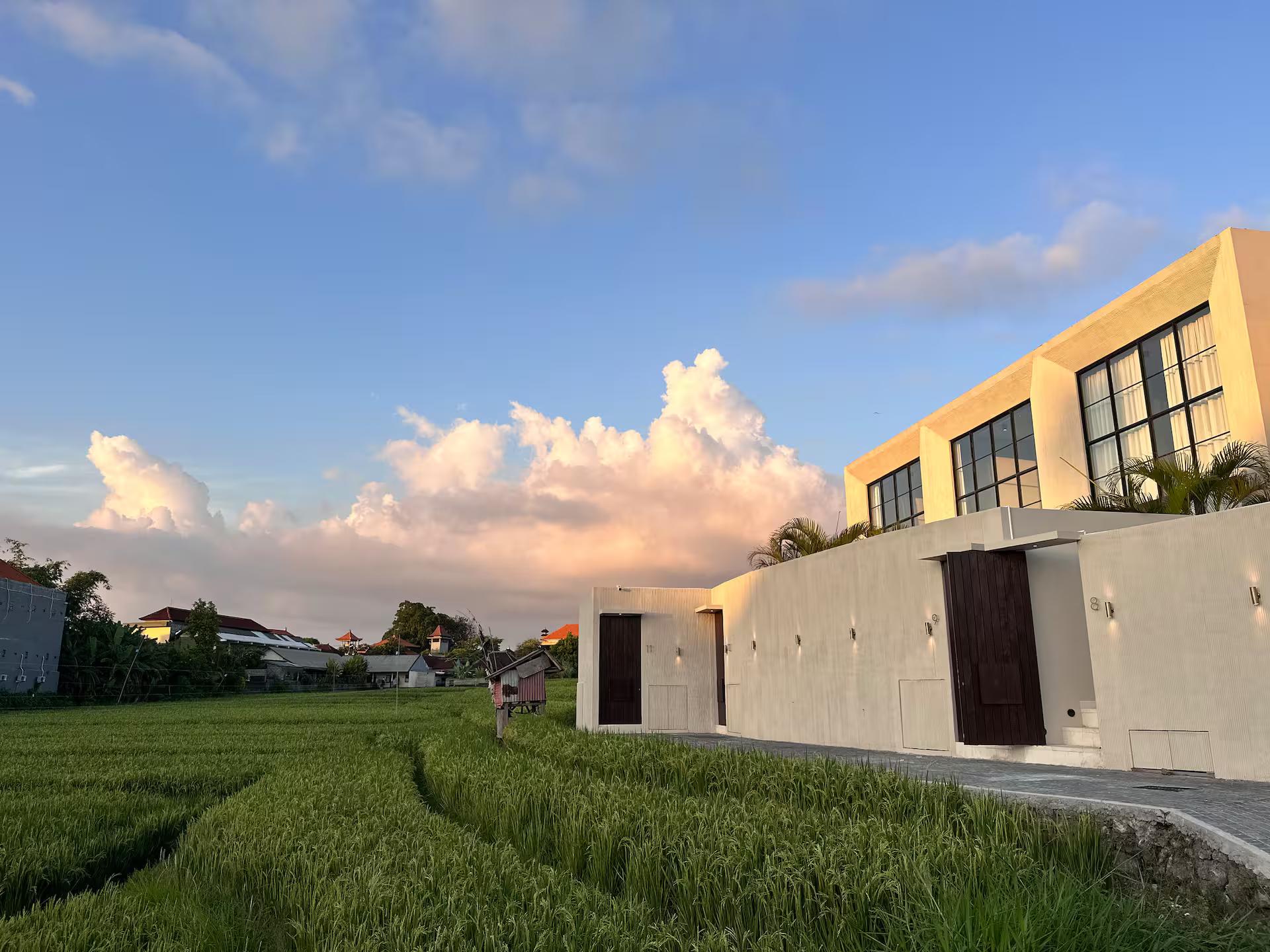 The villa seen across the rice field at dusk, cloud piling up behind the pale render