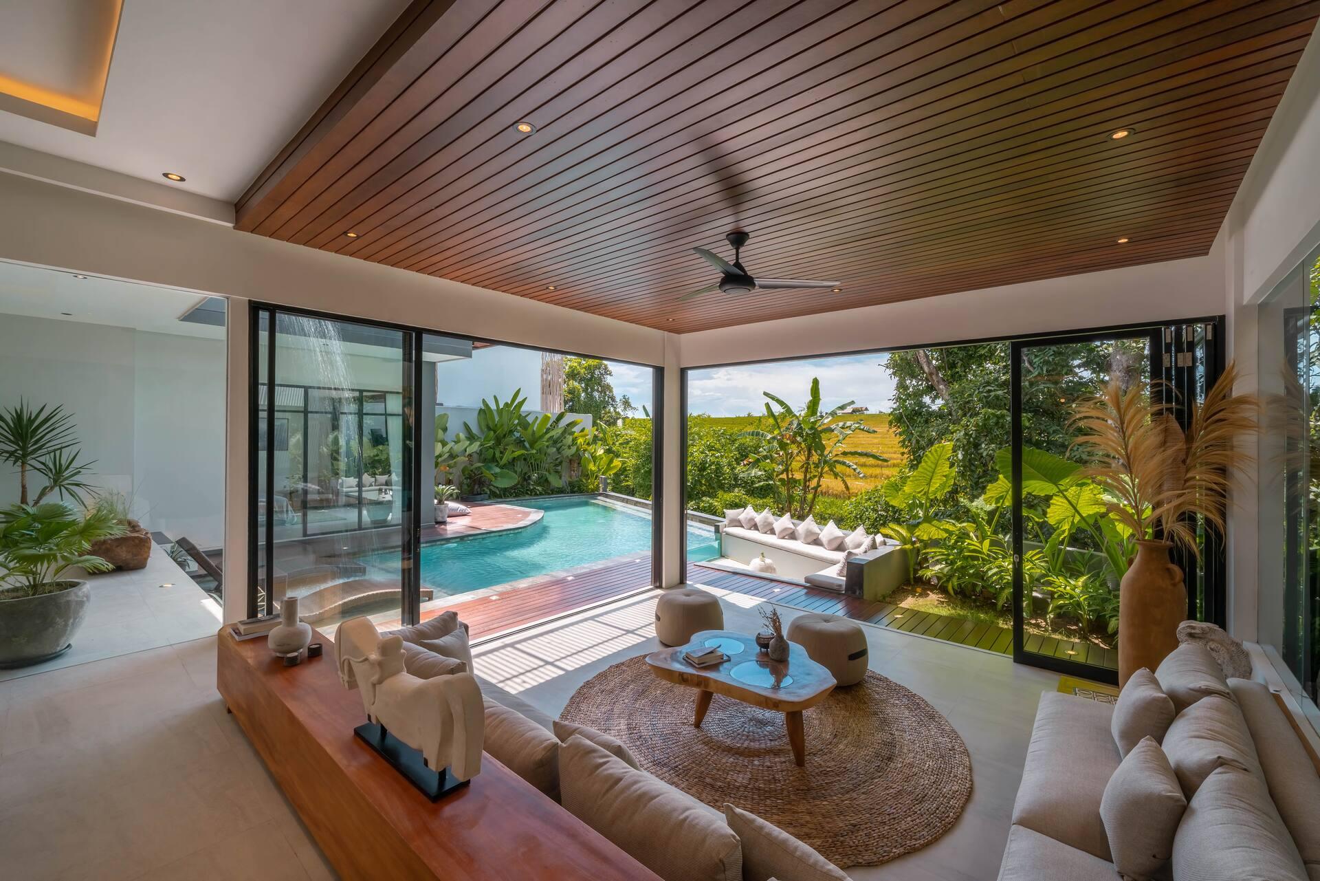 Living pavilion under a dark timber ceiling, open the full width to the pool and the paddies
