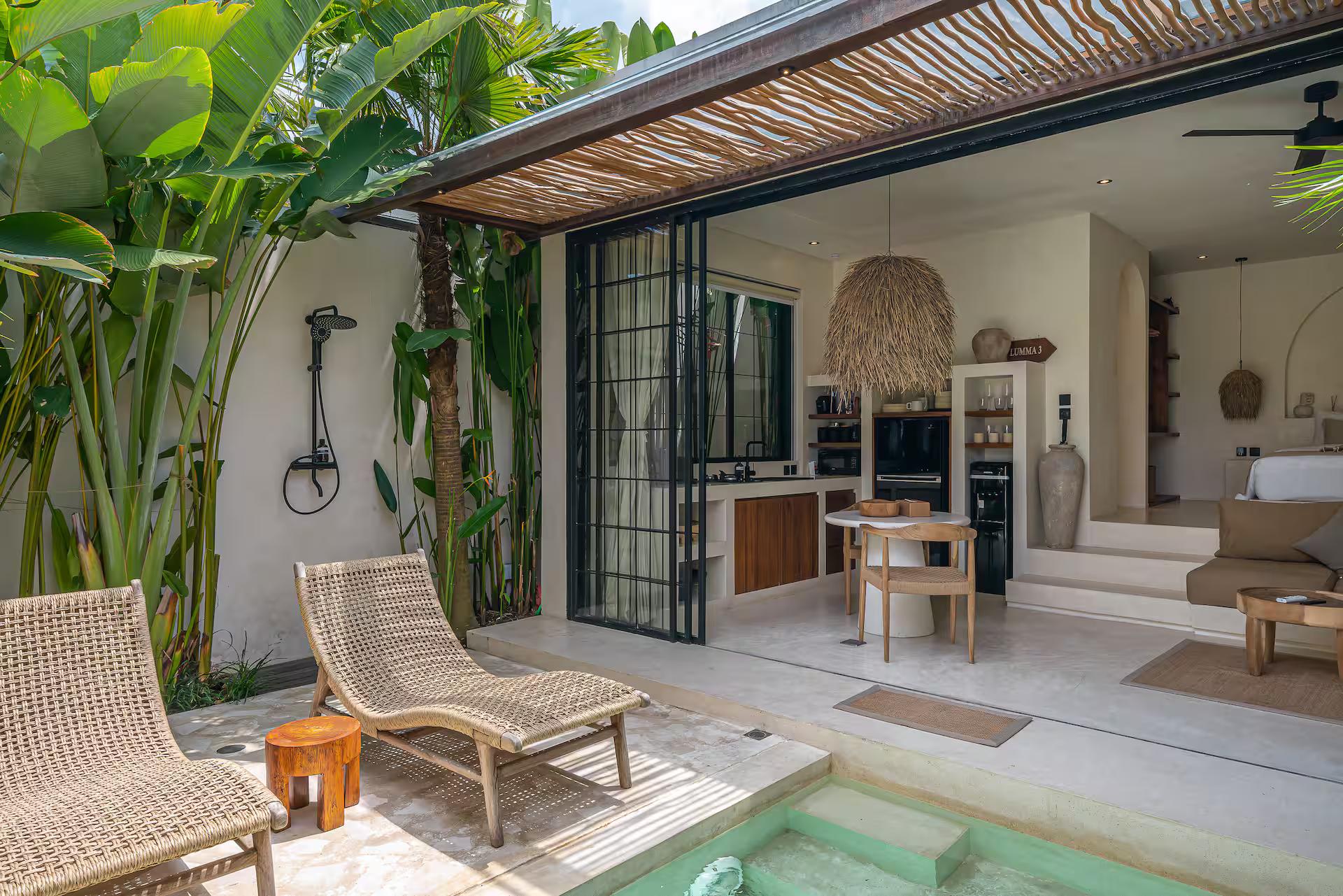 Rattan loungers on the paved terrace beside the pool, glass doors folded open into the pavilion
