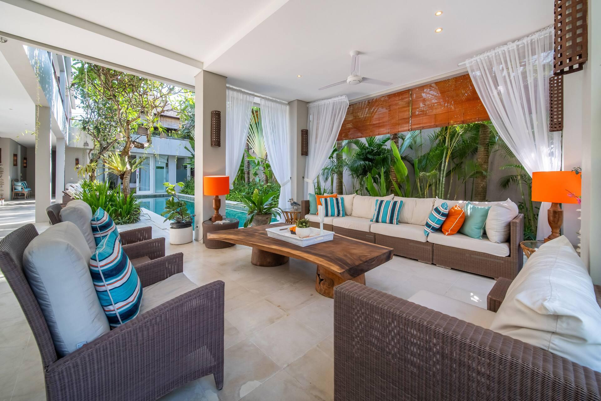 Open-air living room beside the pool, with white drapes, wicker sofas and a carved timber coffee table