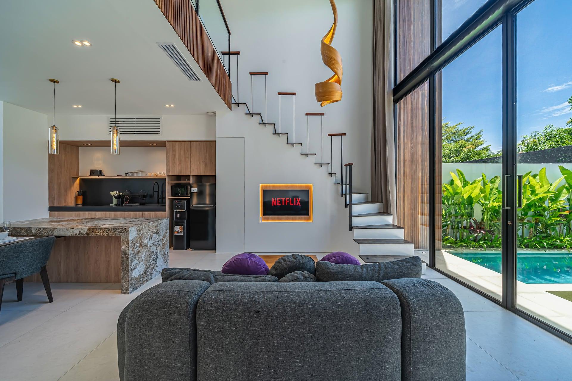 Double-height living room at Aether Villa, with a curved sofa facing the pool doors and the kitchen beyond