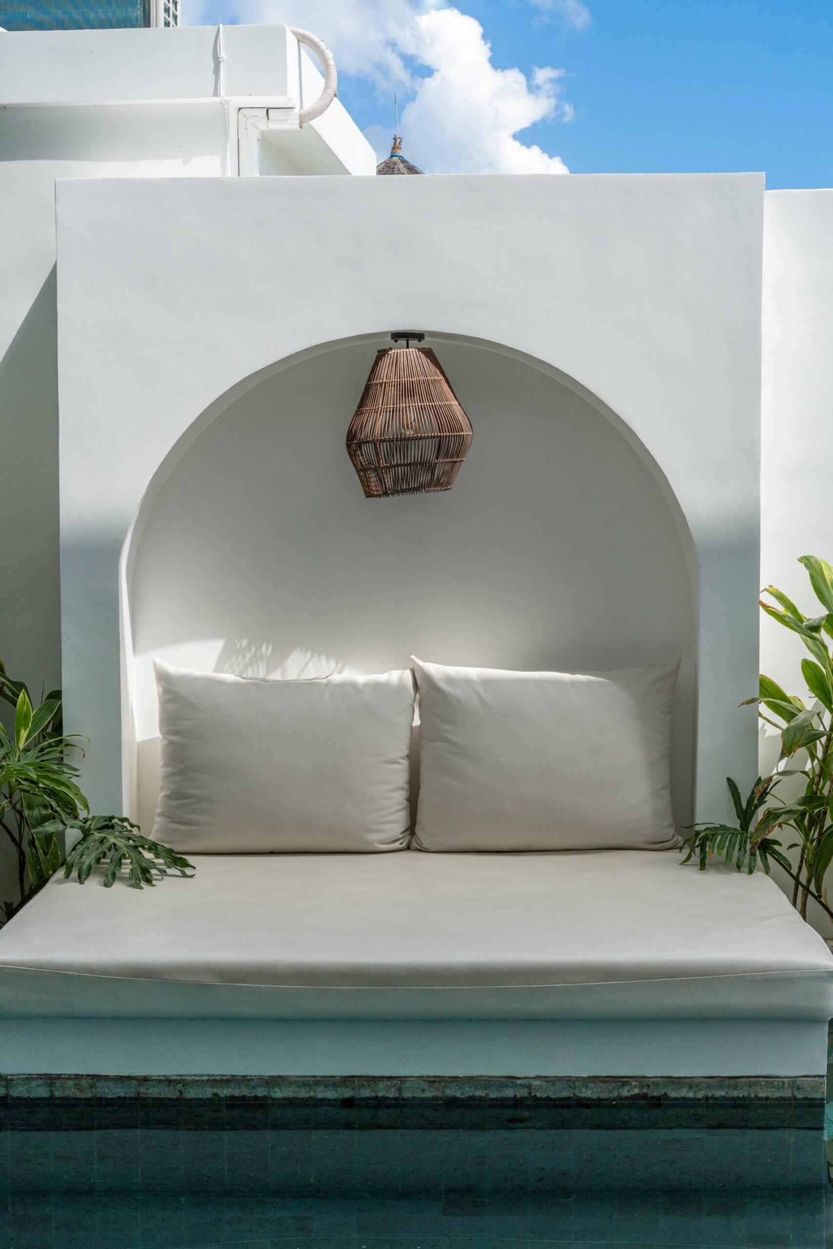 An arched plaster alcove at the head of the pool, cushioned as a shaded daybed under a rattan lantern