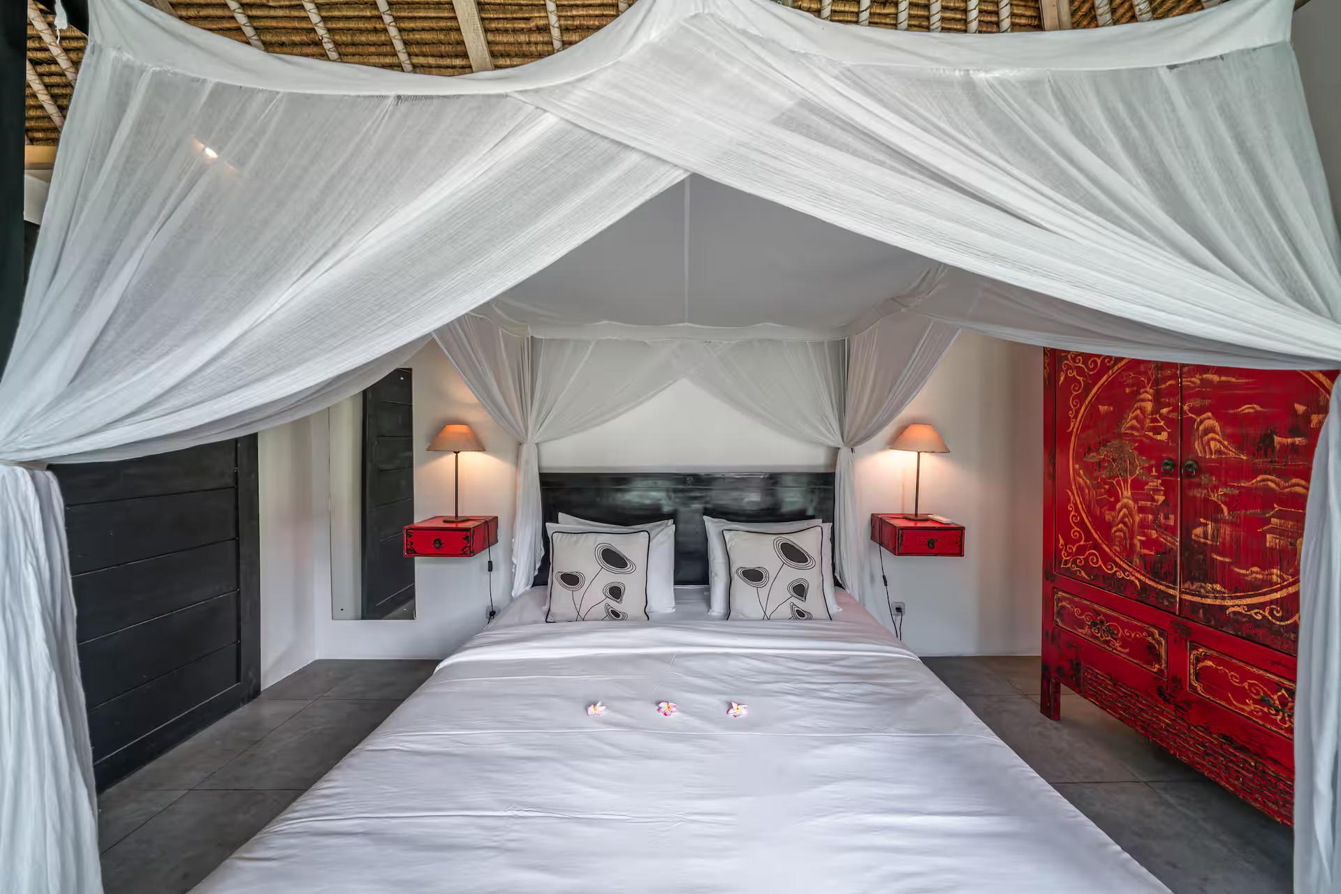 Four-poster bed with a white net and a red lacquered Chinese cabinet alongside