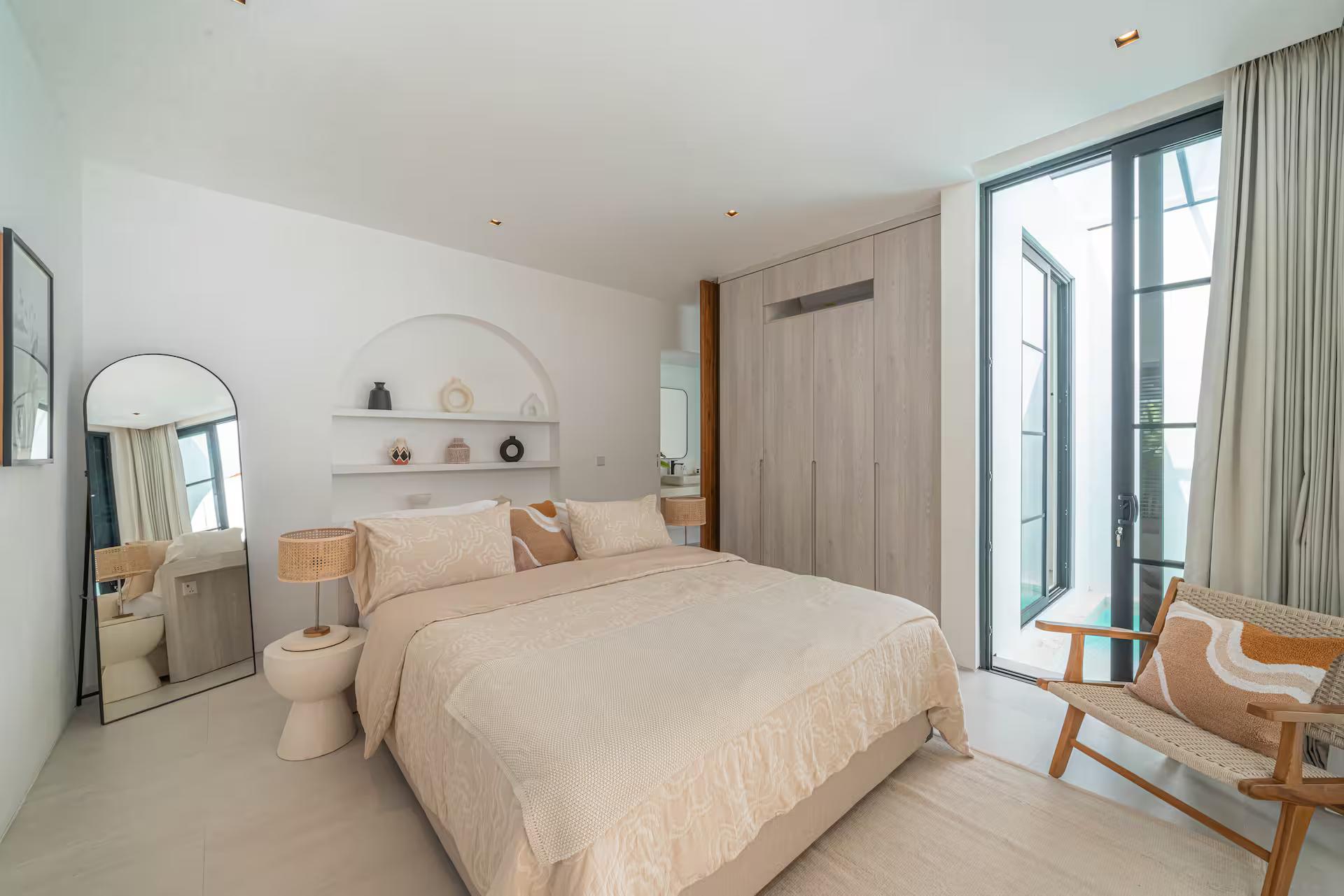 King bedroom with an arched wall niche, leaning mirror and a cane armchair by the window