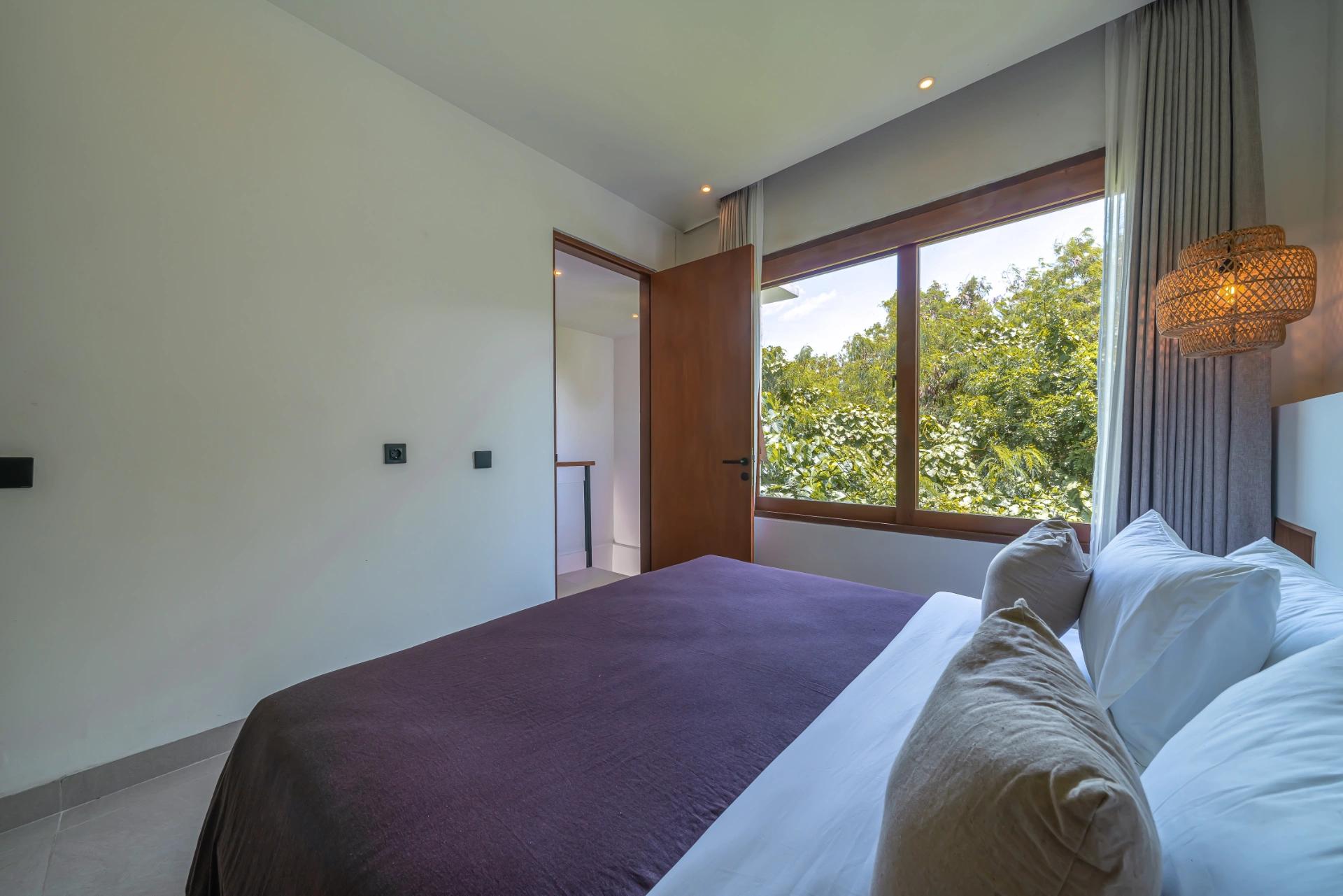 King bed under an aubergine throw beside a picture window looking into the treetops