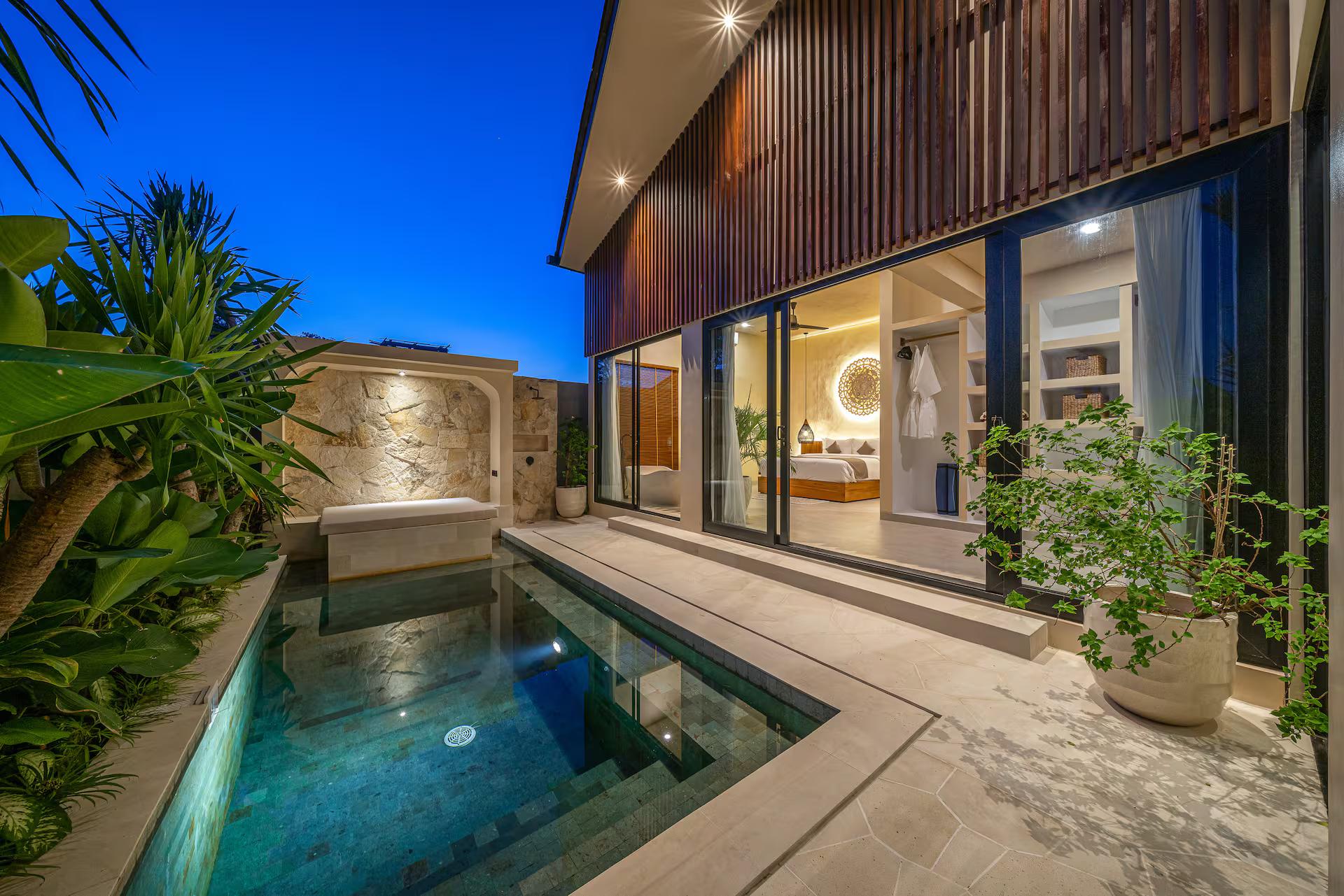 Villa Seabel at dusk, the pool lit beside a stone wall with an arched recess and built-in daybed