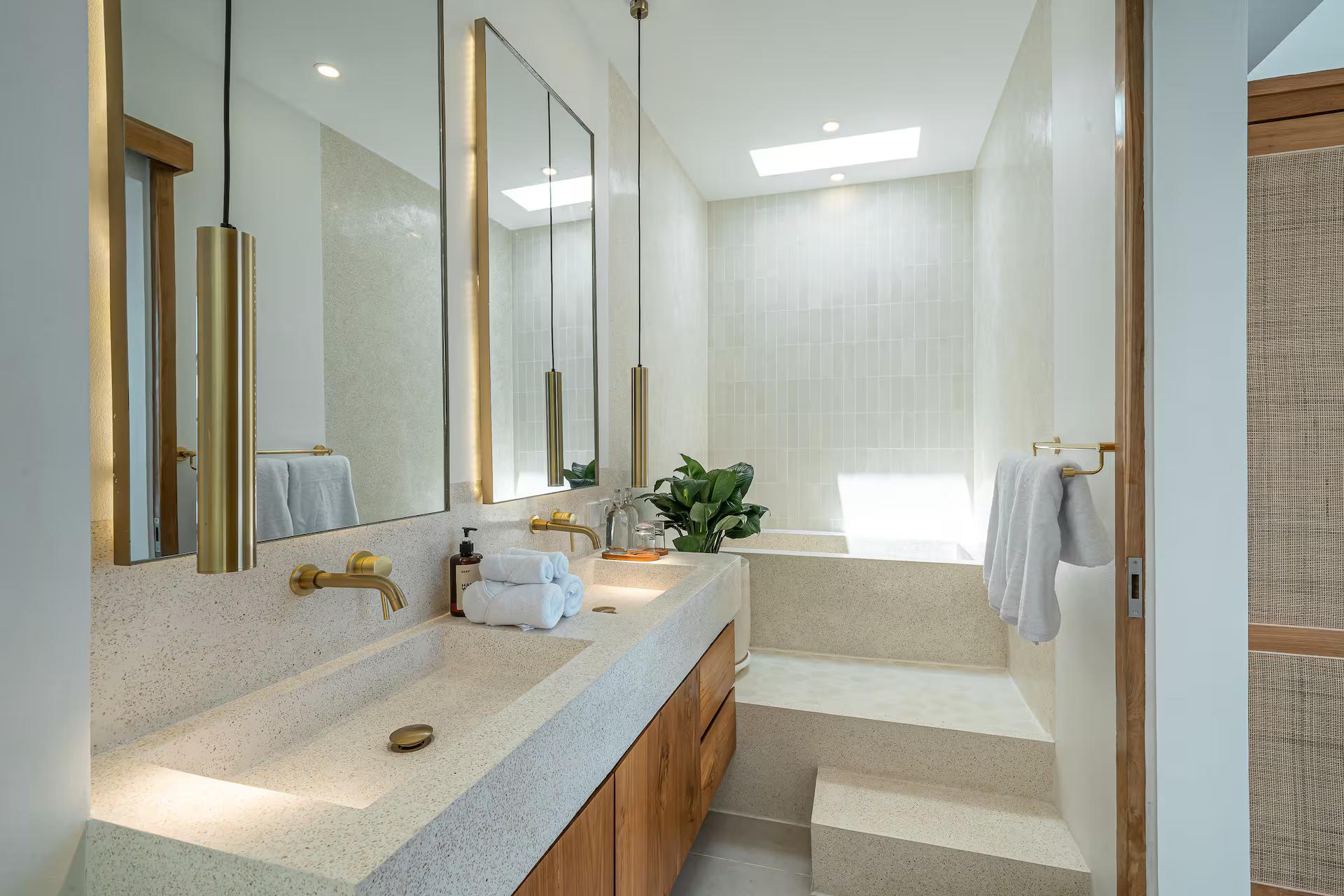Terrazzo trough basin with brass wall taps and a sunken tub under a skylight