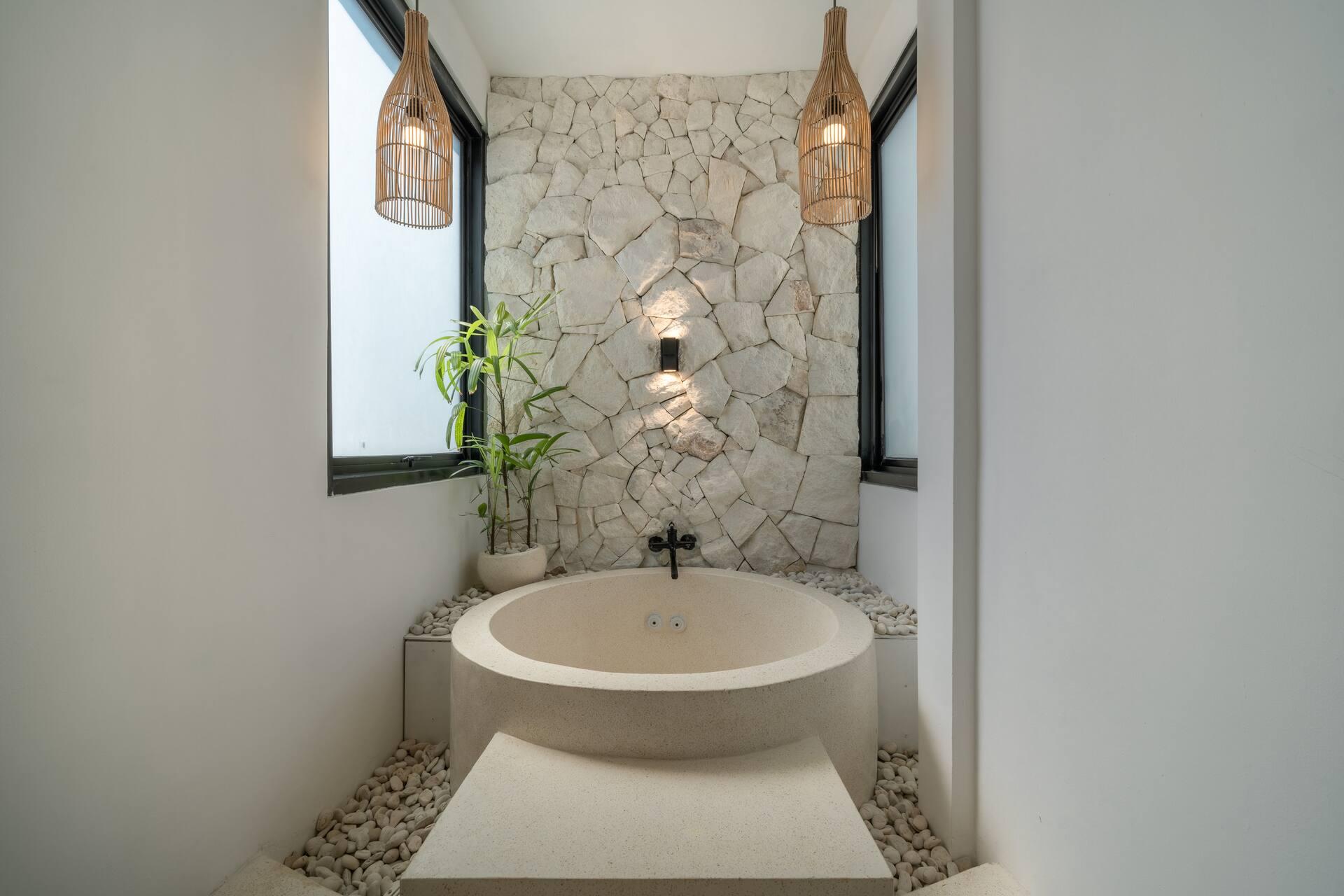 Round stone soaking tub on white pebbles beneath a stone-clad wall and tall window in the ensuite