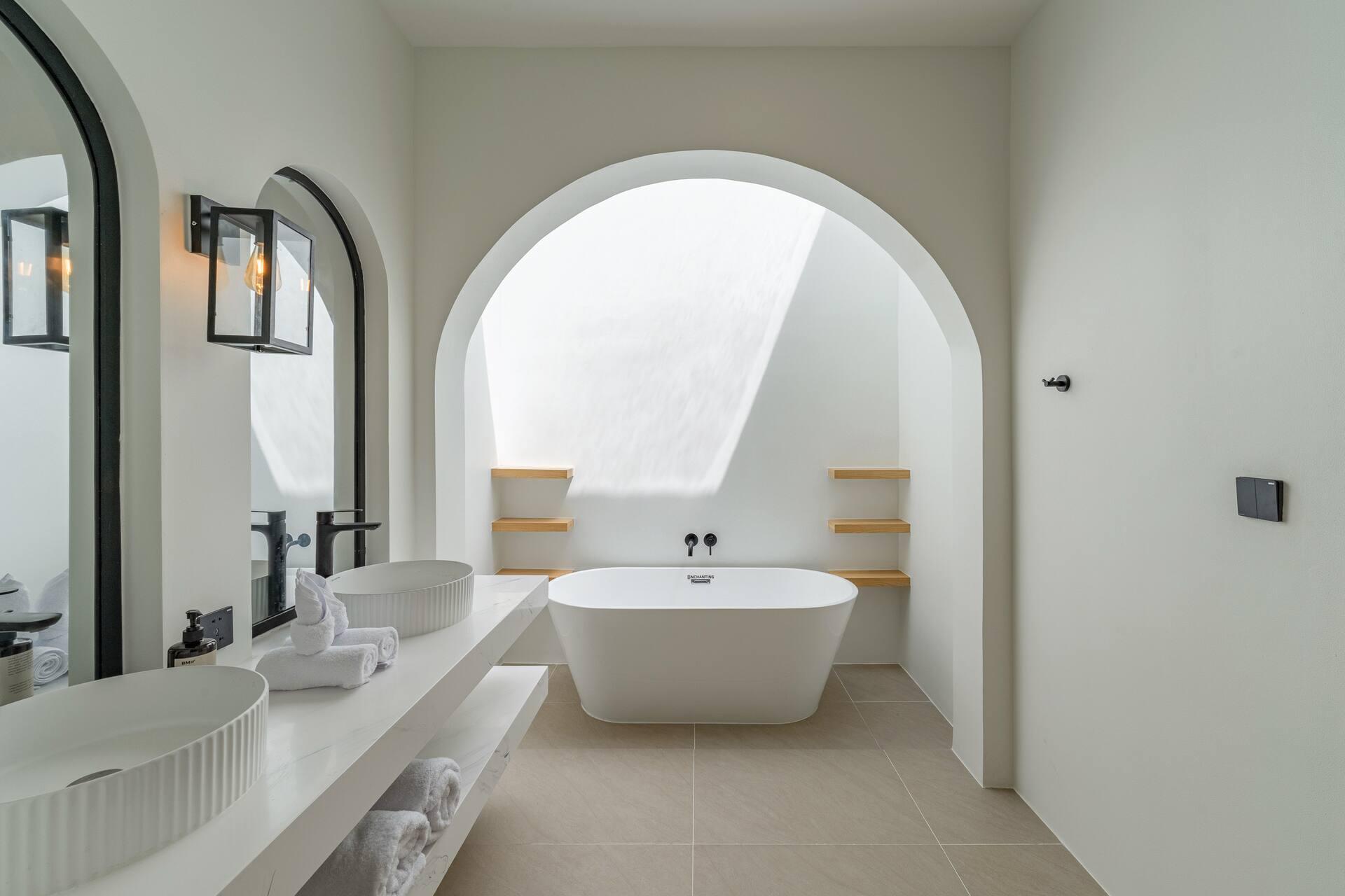 Ensuite with a freestanding bath set in a skylit arch and a white double vanity with black wall lamps