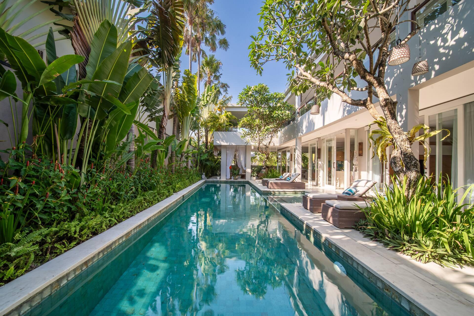 The long turquoise pool framed by tropical planting, loungers and a thatched gazebo at the far end