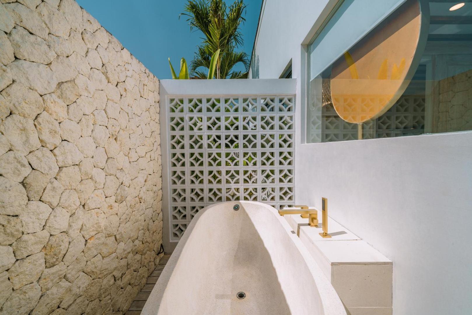 Outdoor bathtub in white plaster with brass tapware against a rough coral-stone wall