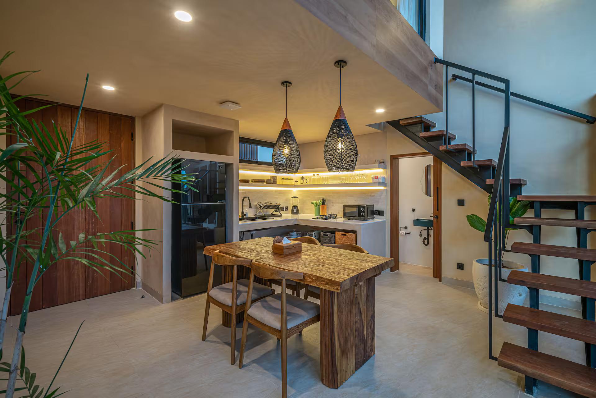 Live-edge suar dining table under black rattan pendants, the open-tread stair rising behind