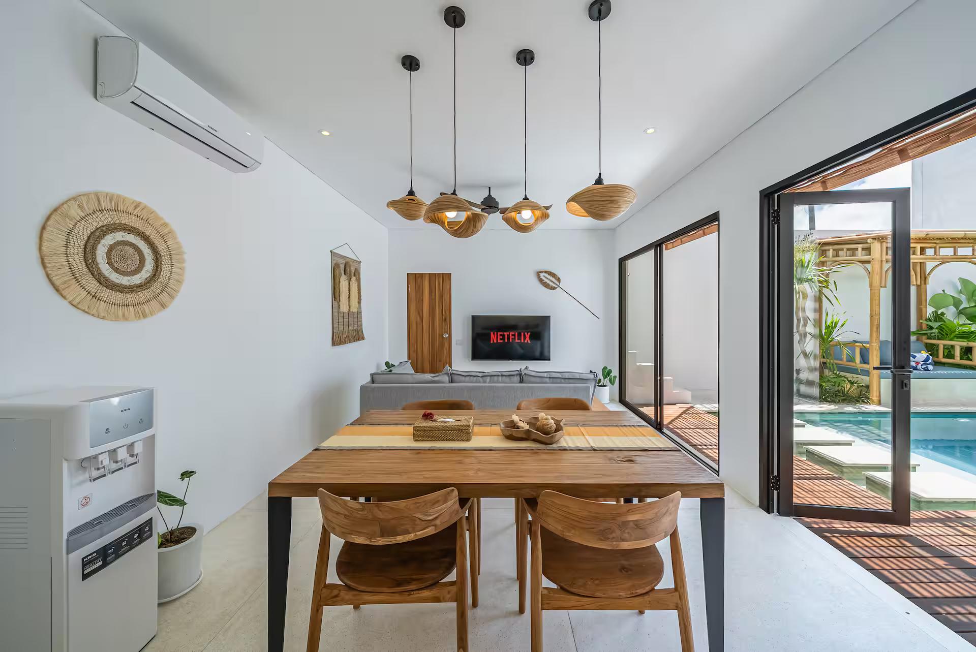 Live-edge timber dining table under woven rattan pendants, pool deck through the glass