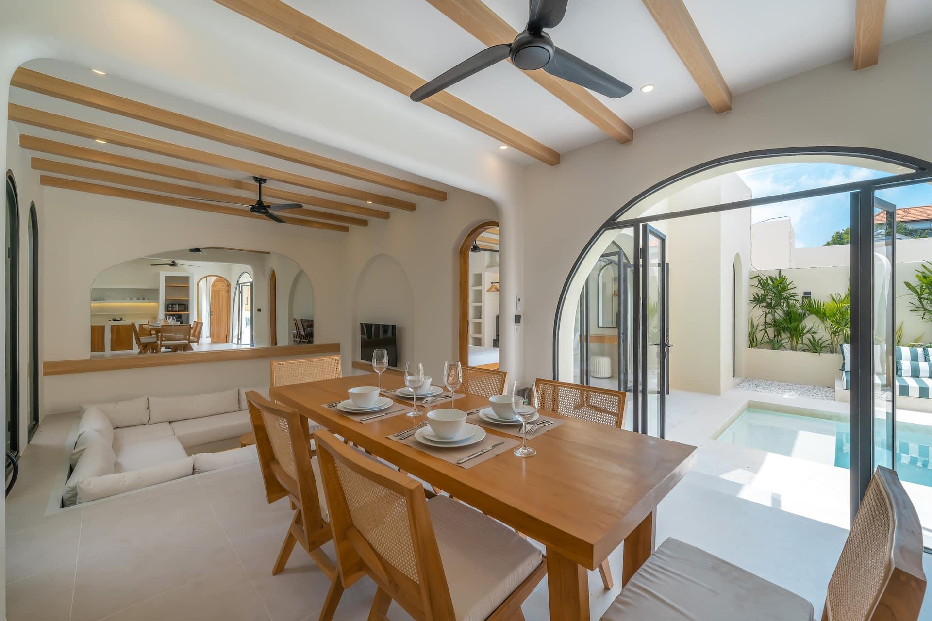 The oak dining table laid for six, the pool visible through the arched pivot doors beyond