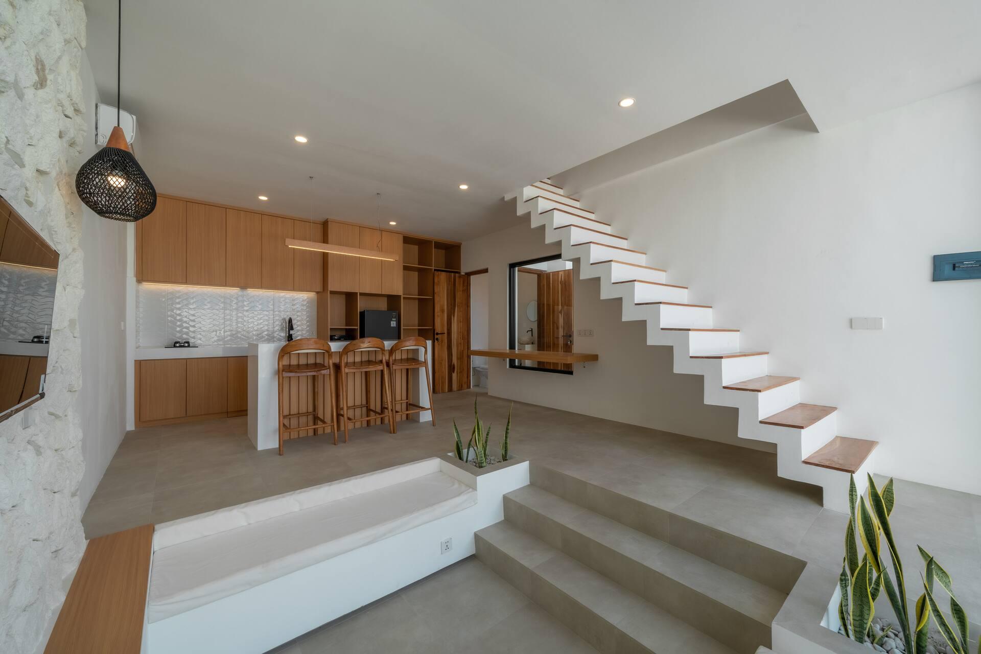 Open-plan living area with a floating white staircase, timber kitchen and bar stools at Villa Merine