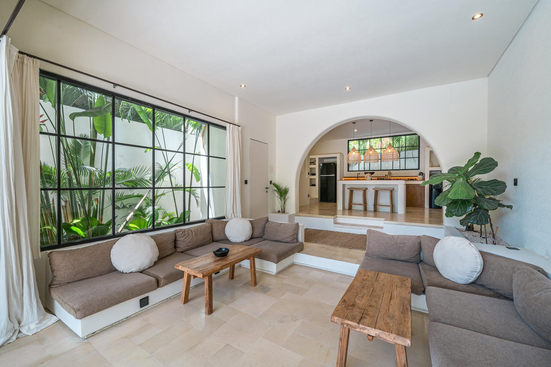 Living room at Villa Ania with tall black-framed windows onto tropical planting and the arched kitchen beyond