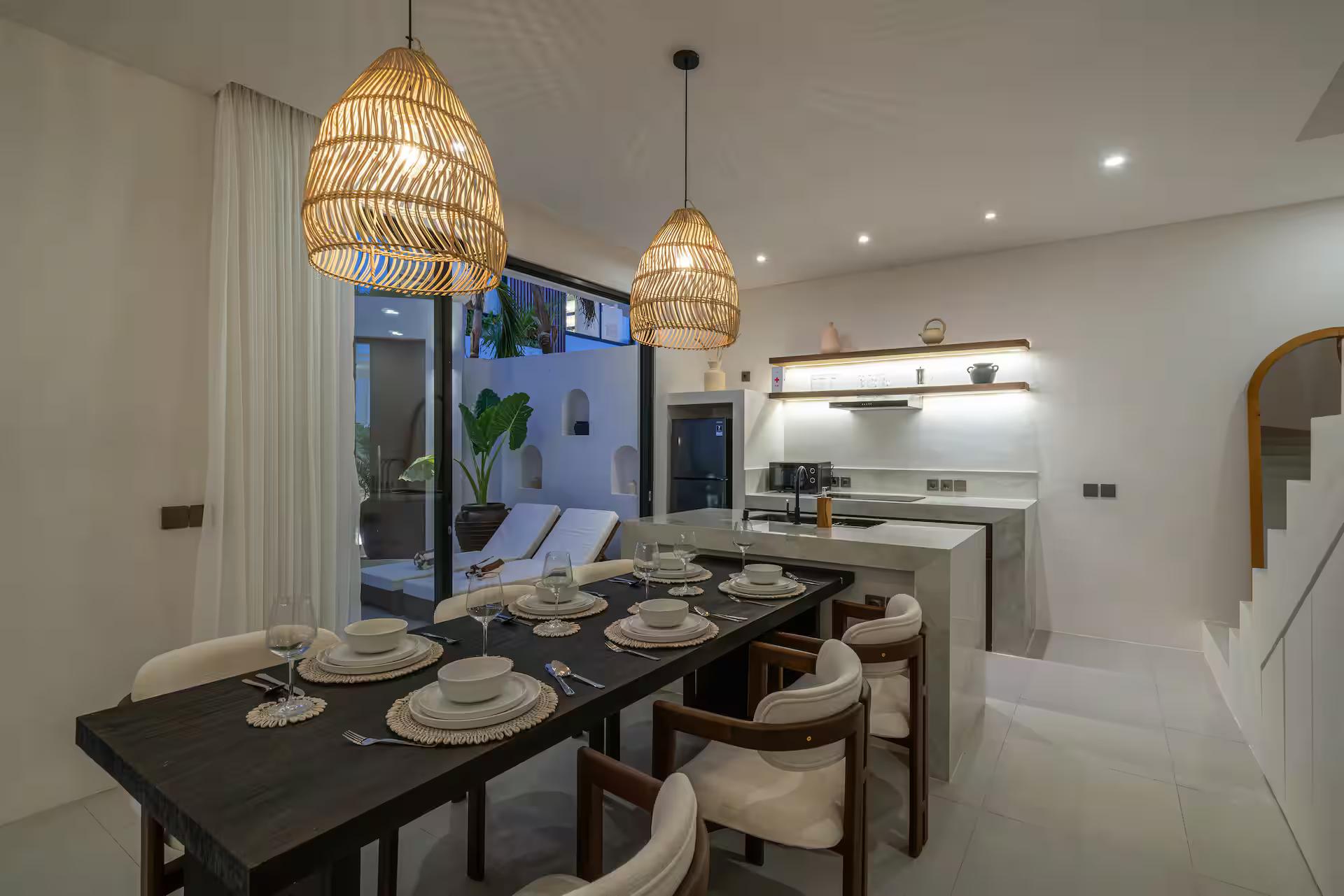 Dark timber dining table laid for six under three rattan pendants beside the kitchen