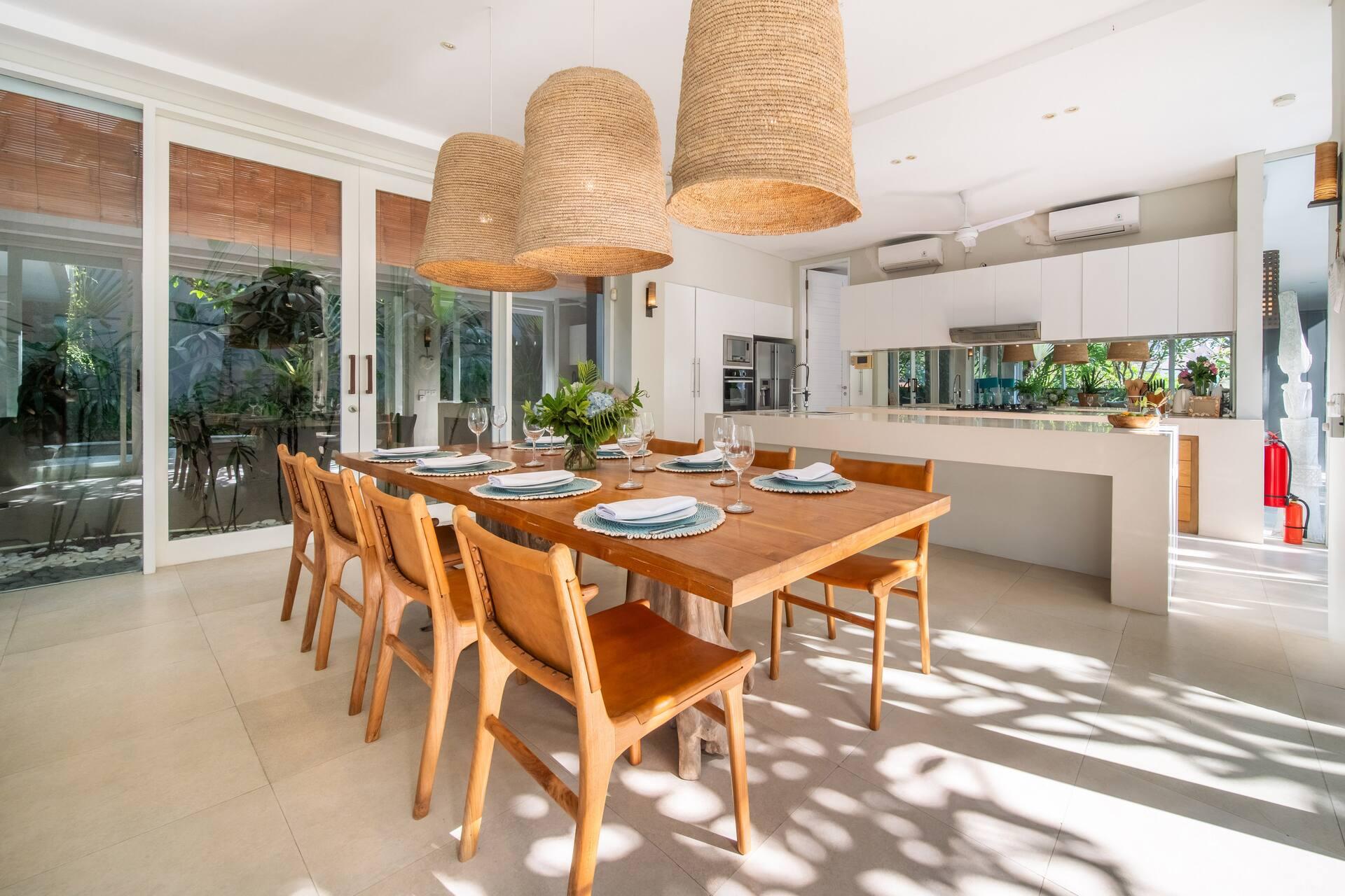 Teak dining table for ten under three large woven pendants, with the open kitchen alongside