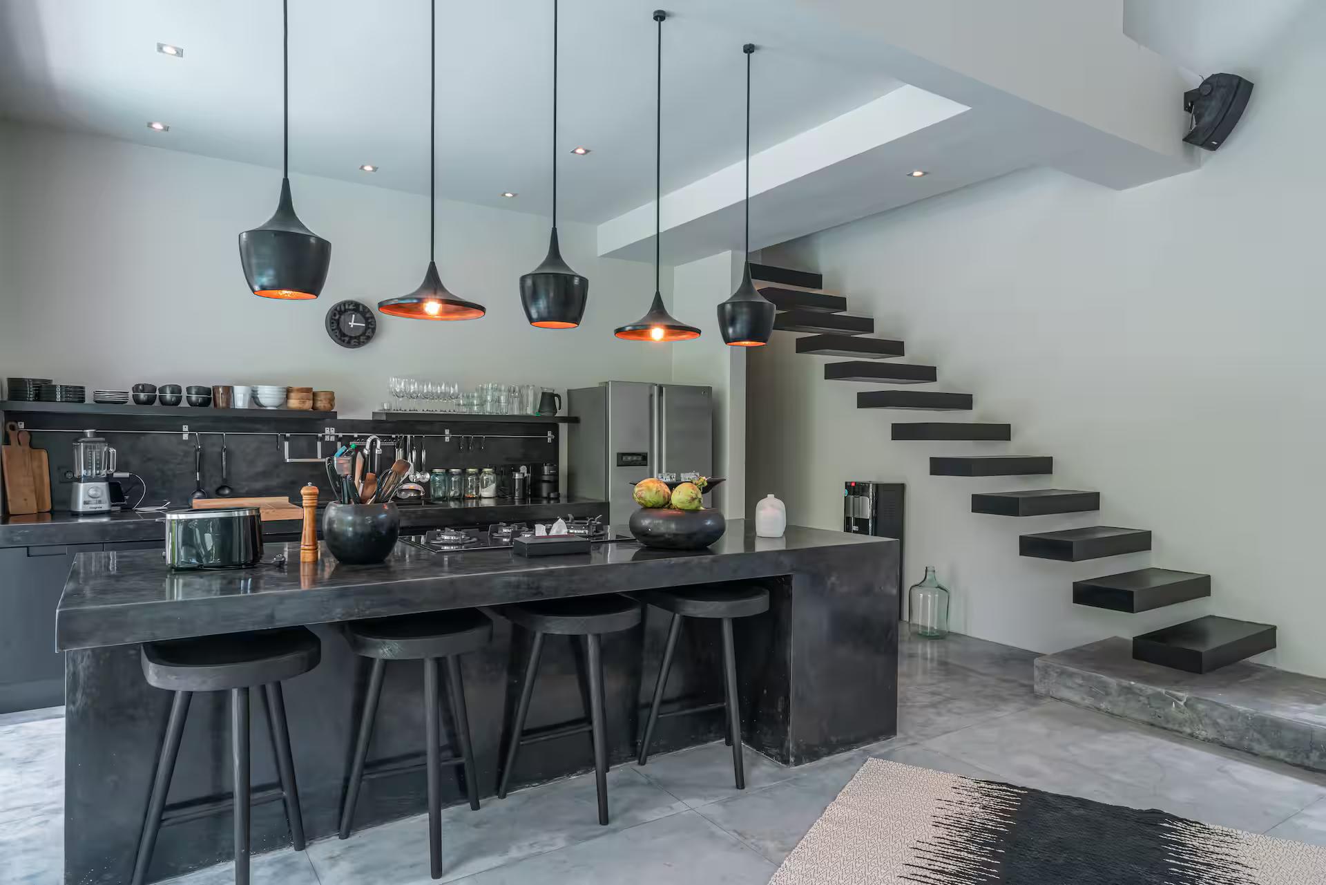 Black concrete kitchen island with bar stools and copper pendants below the floating stair