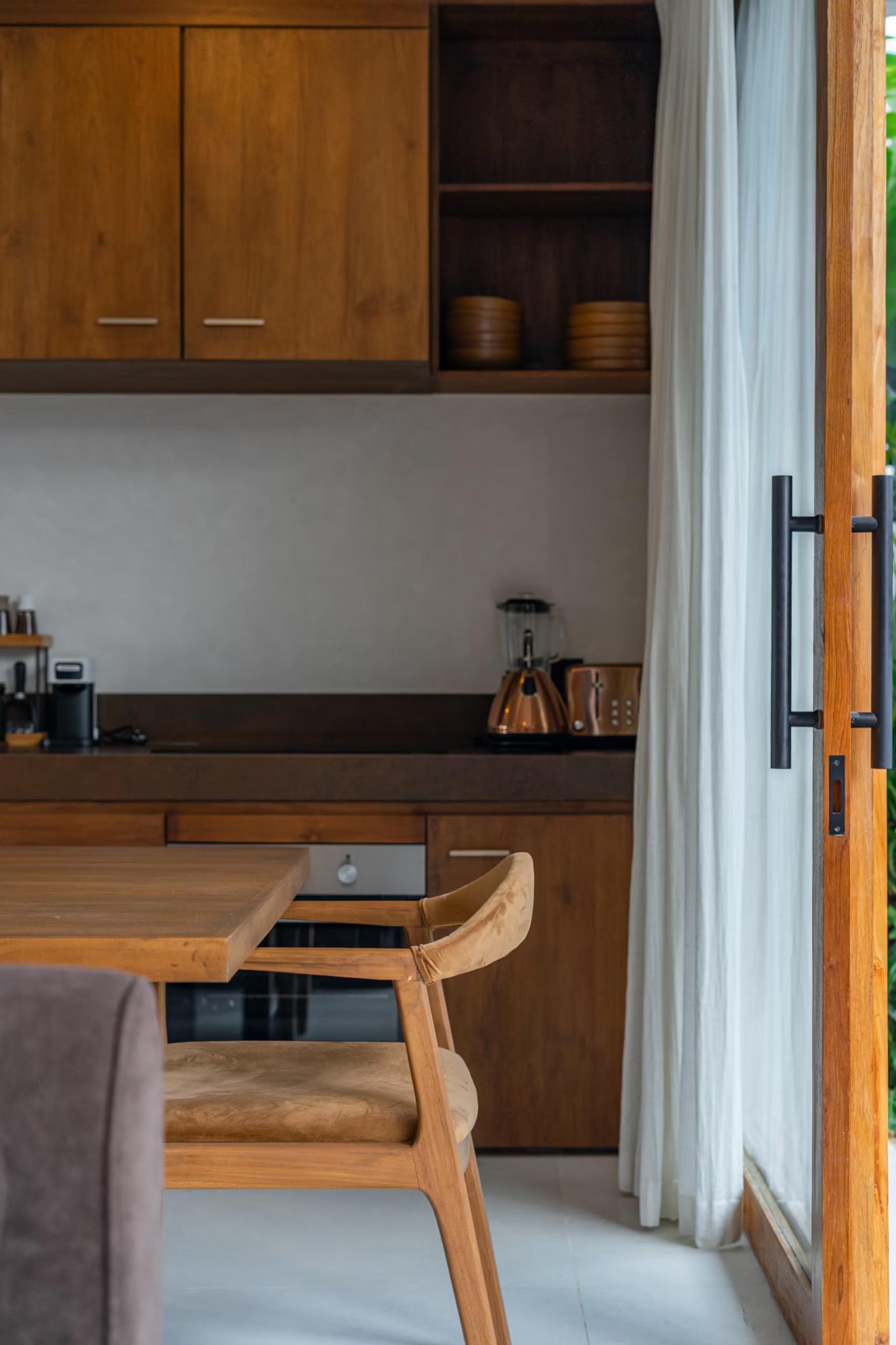 Teak dining table and woven-seat chairs beside the kitchen joinery, curtains drawn back to the terrace