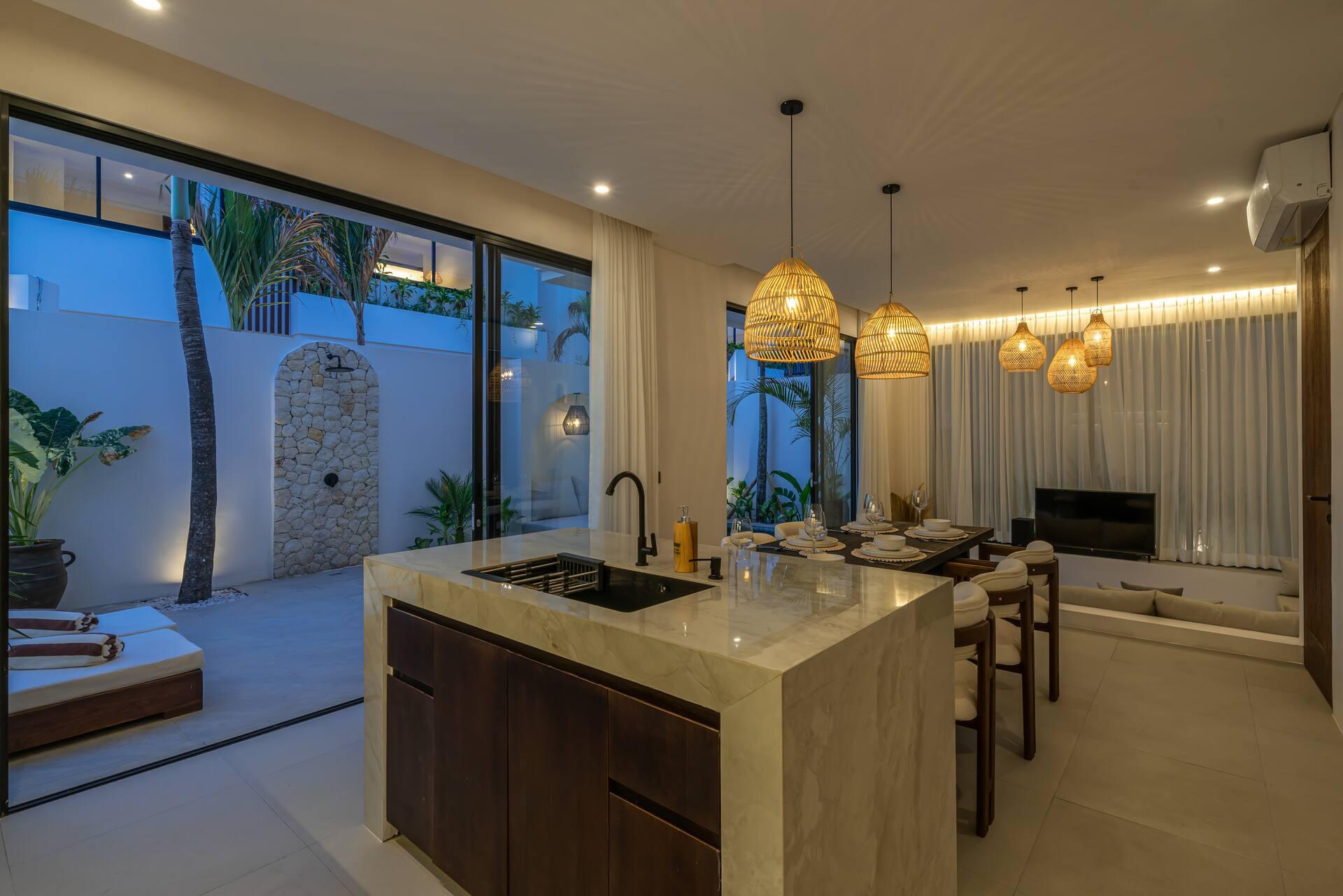 Open-plan kitchen and lounge at Villa Nyra under rattan pendants, with the stone shower wall outside