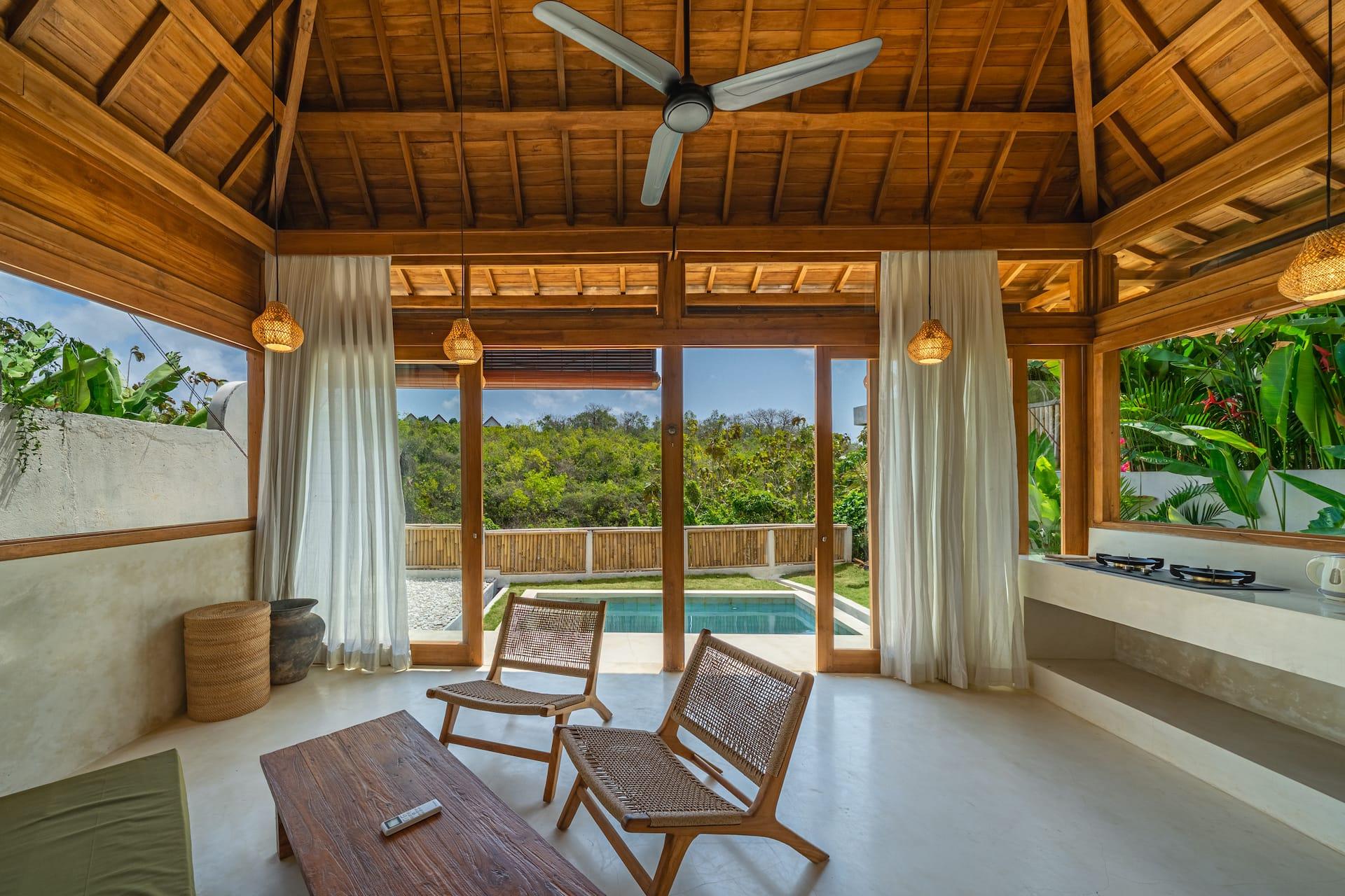 Living pavilion with woven lounge chairs and sheer curtains opening onto the pool and greenery