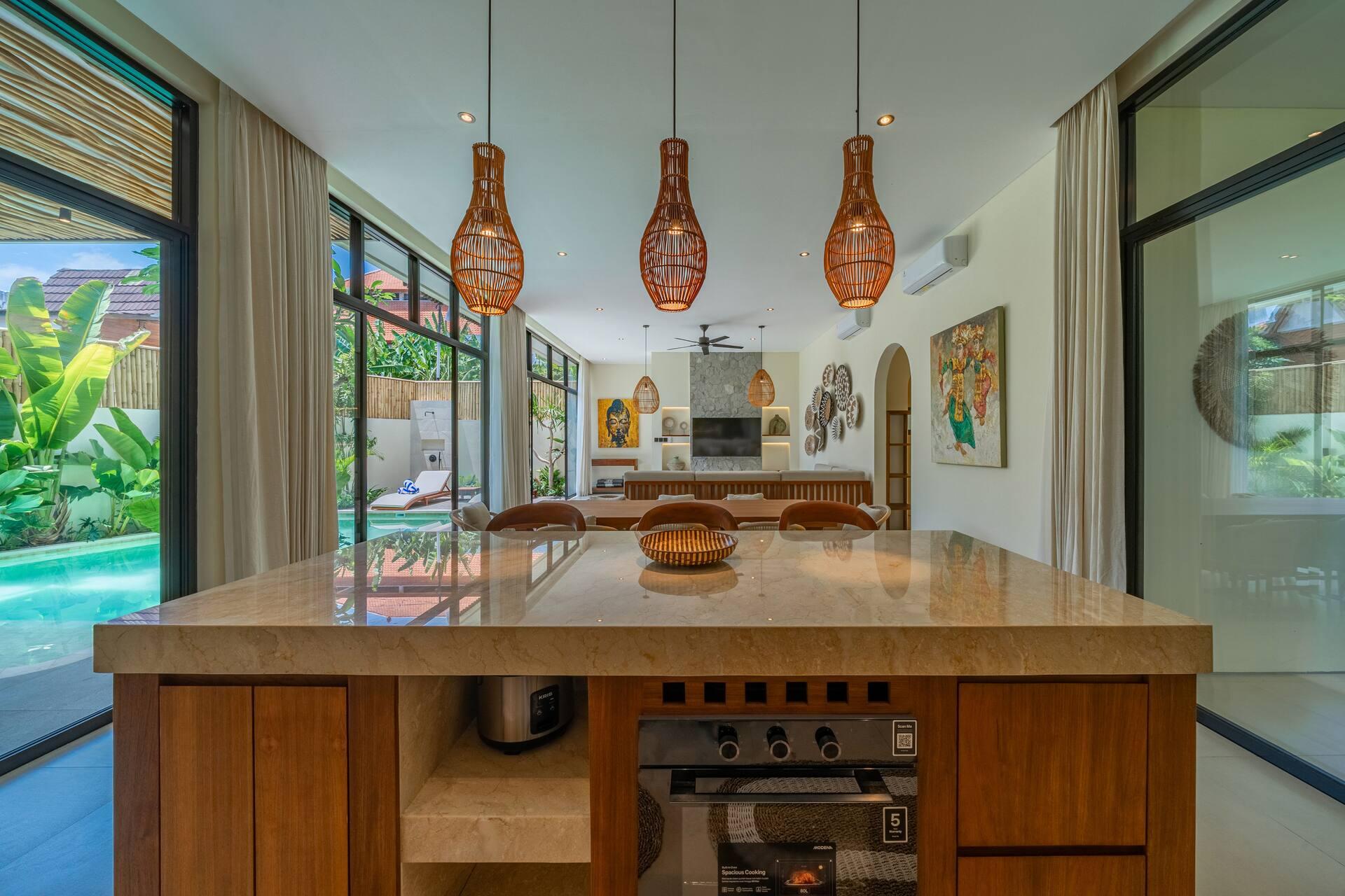 Kitchen island with a stone top and oven, rattan pendants overhead and the pool beyond the glass