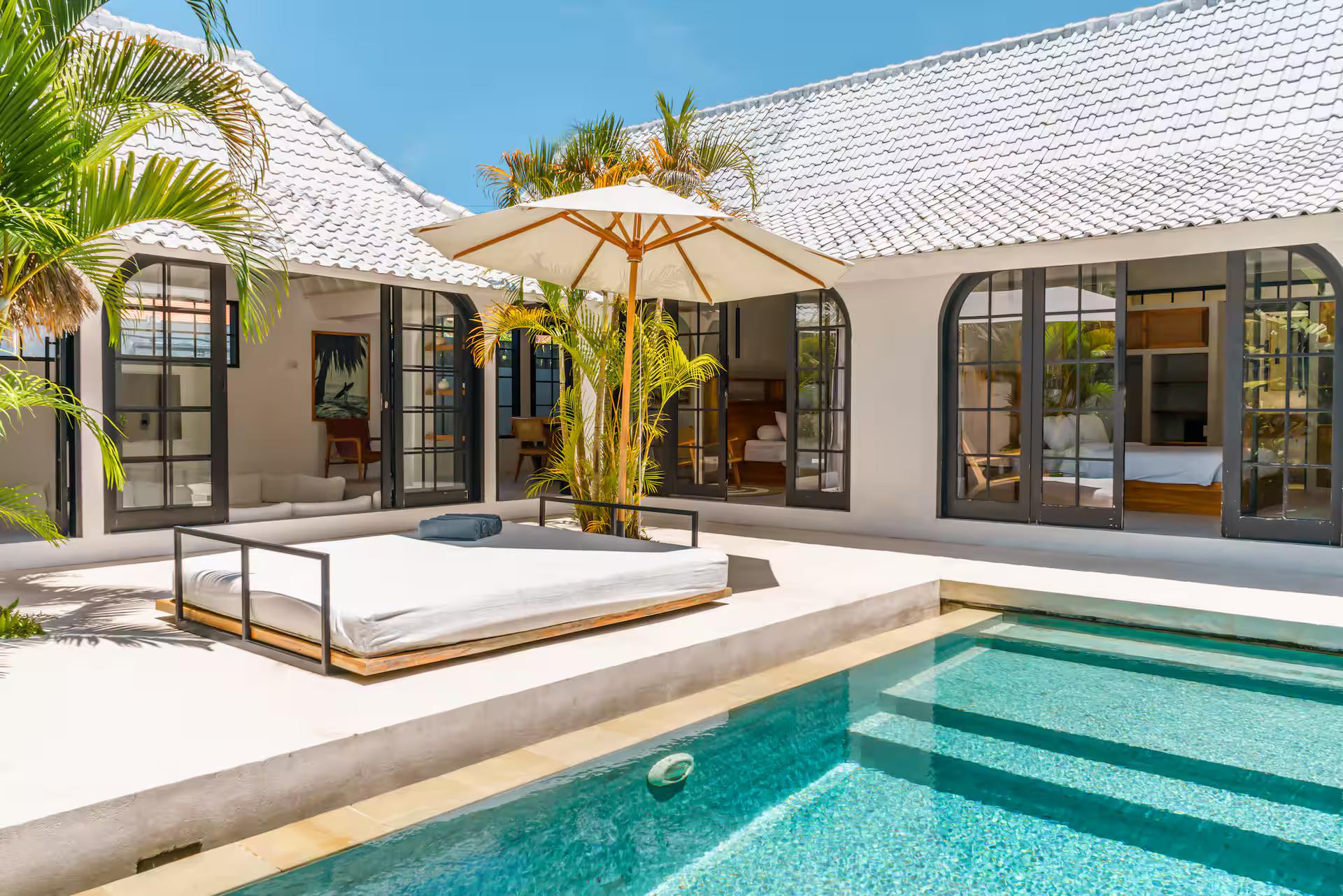 Timber-framed daybed under a canvas parasol at the head of the pool, arched steel doors behind
