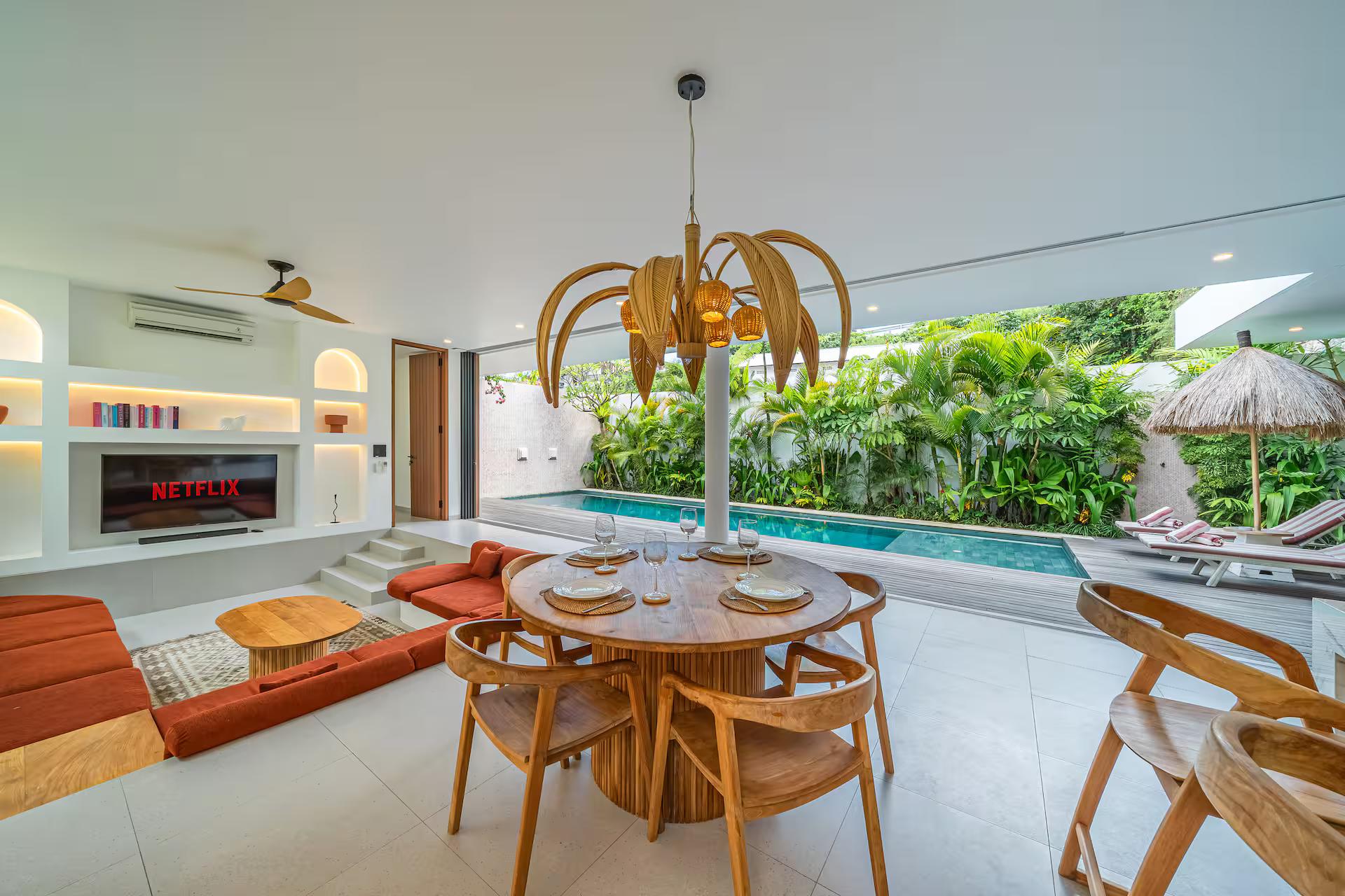 Round timber dining table and rattan chairs under a brass palm-frond chandelier, open to the pool