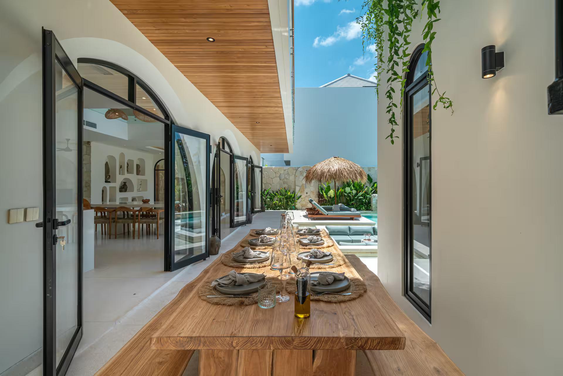 Long timber table laid for dinner on the covered terrace, arched glass doors folded back