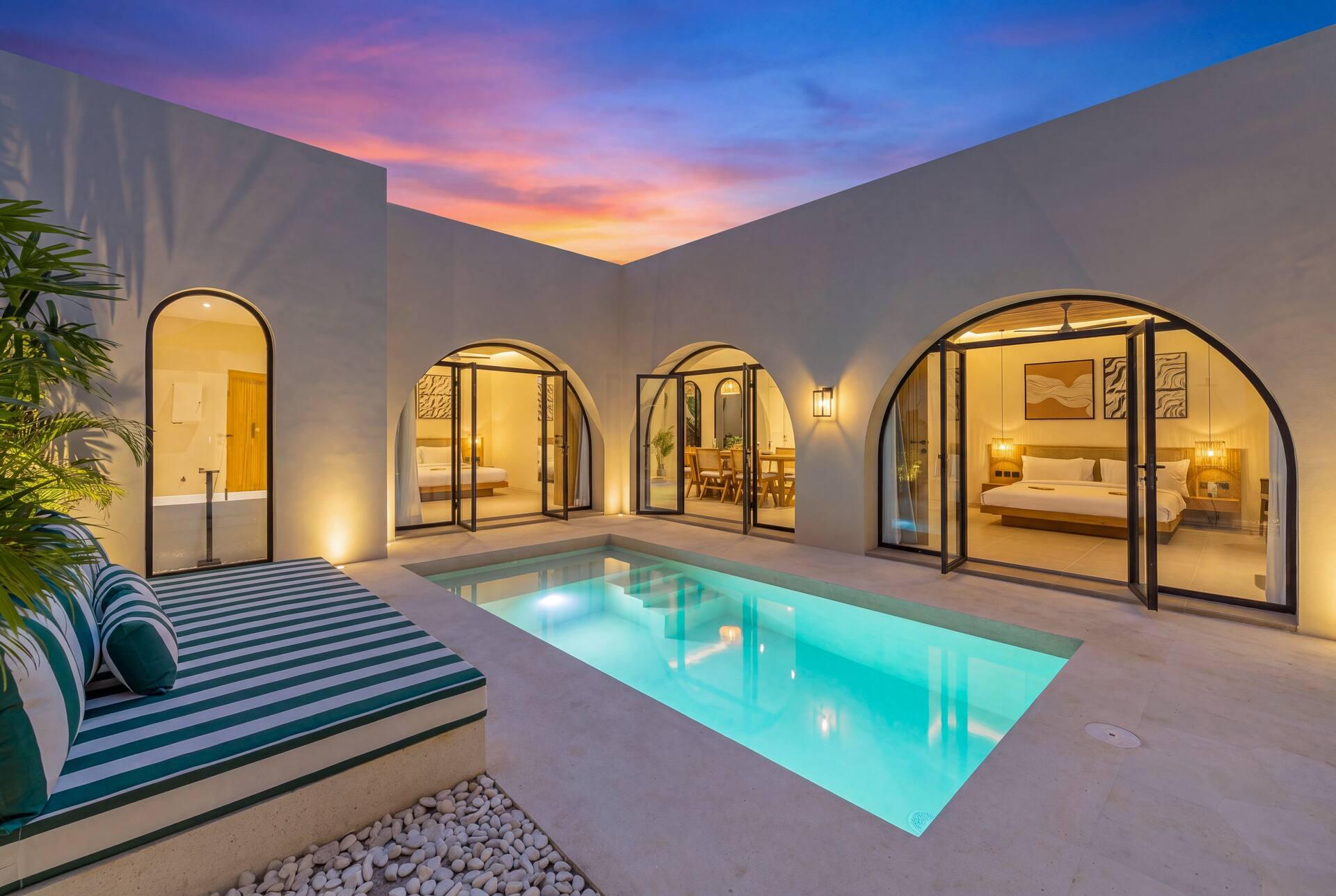 The pool courtyard at dusk, lit arched doors glowing around the water and a green-and-white striped daybed