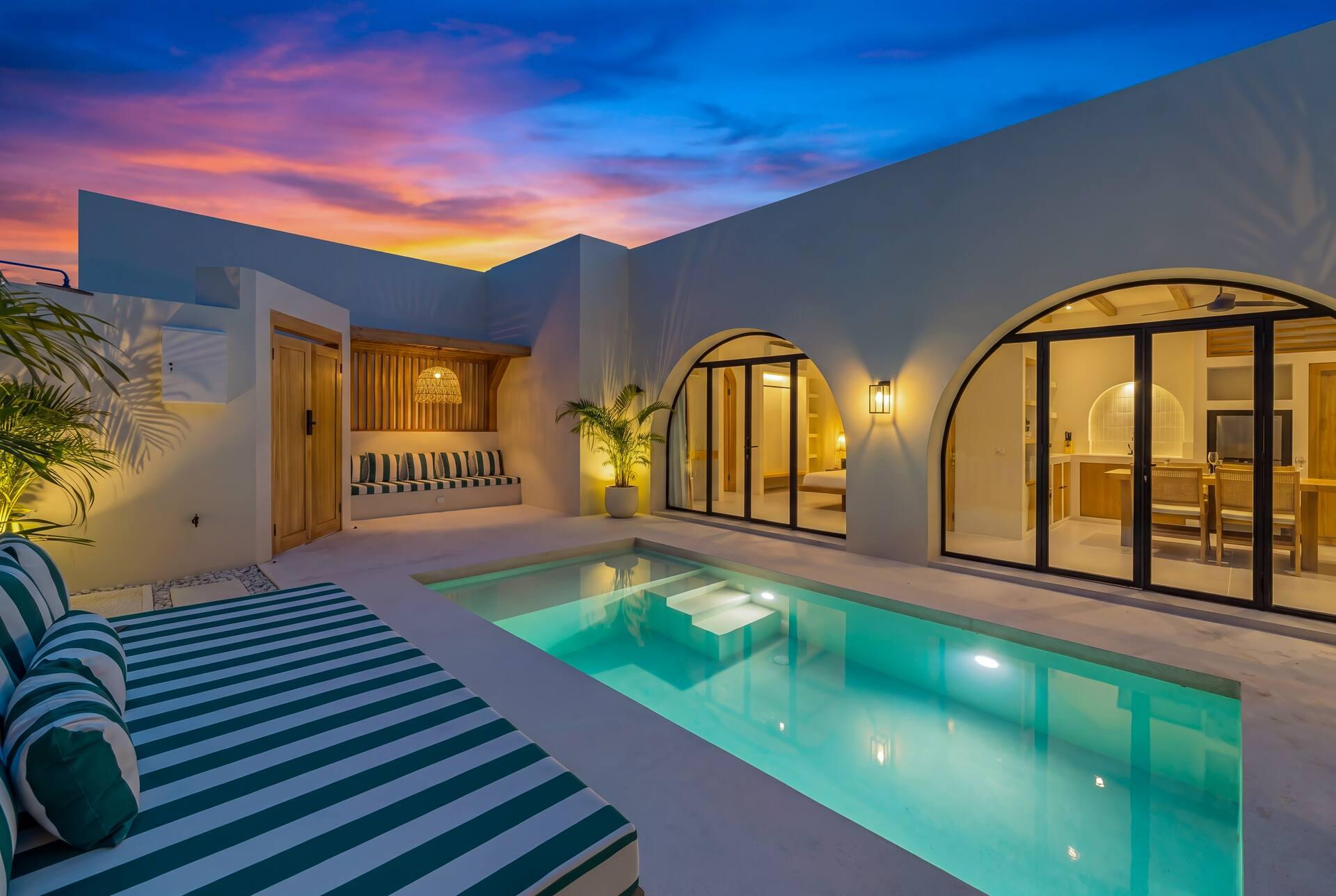 The pool courtyard at sunset, lit arches glowing and a green-and-white striped lounger at the water's edge