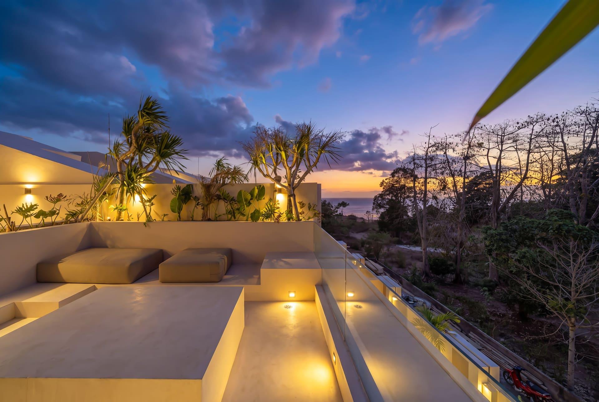 Rooftop lounge at sunset, low built-in seating lit from beneath and the sea on the horizon beyond the trees