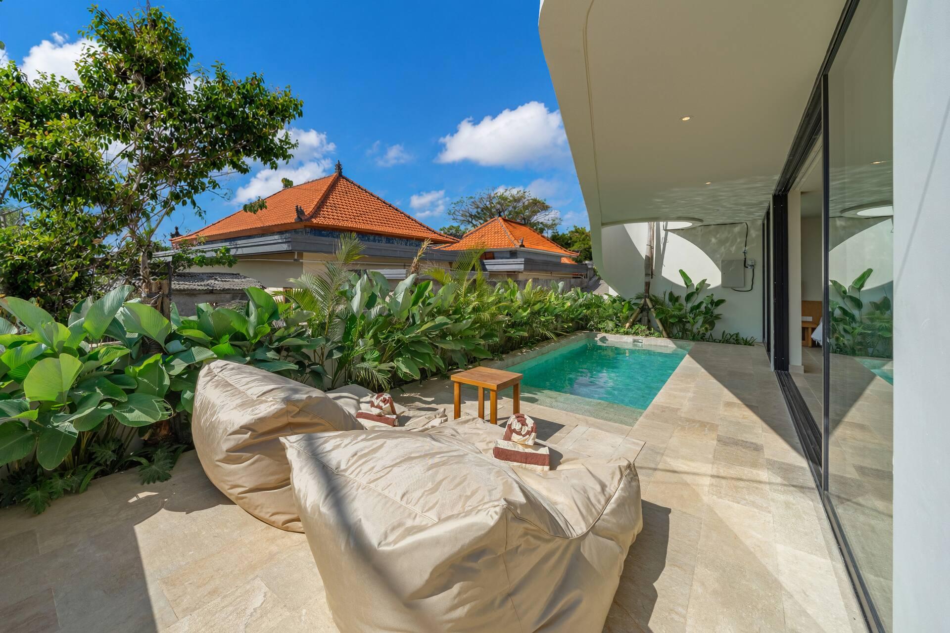 Bean-bag lounger and a low teak table on Villa Aura's pool deck, a temple roof beyond the wall