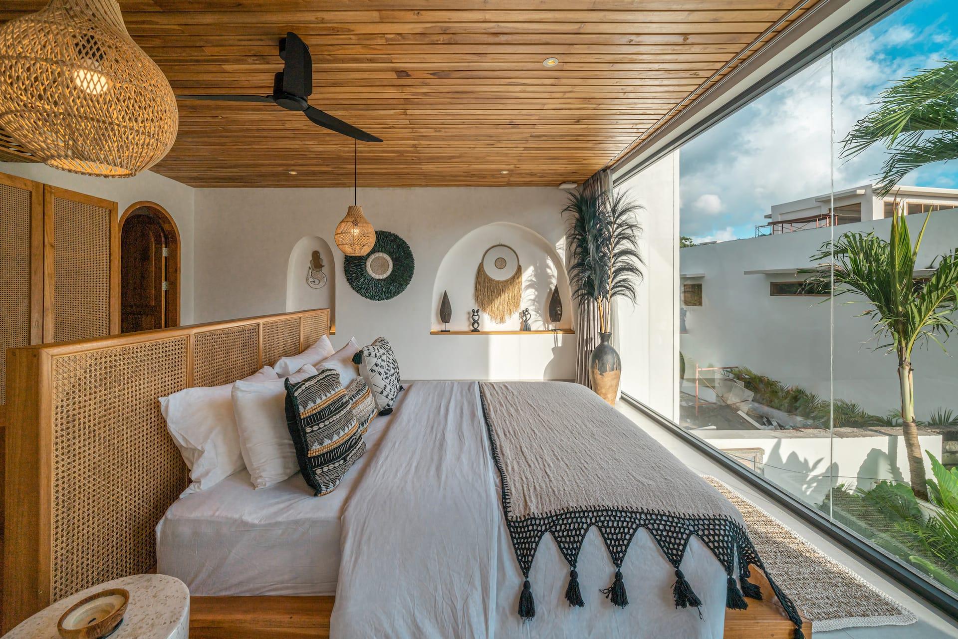 Villa Dharma bedroom with a woven headboard, tasselled throw and floor-to-ceiling glass onto palms
