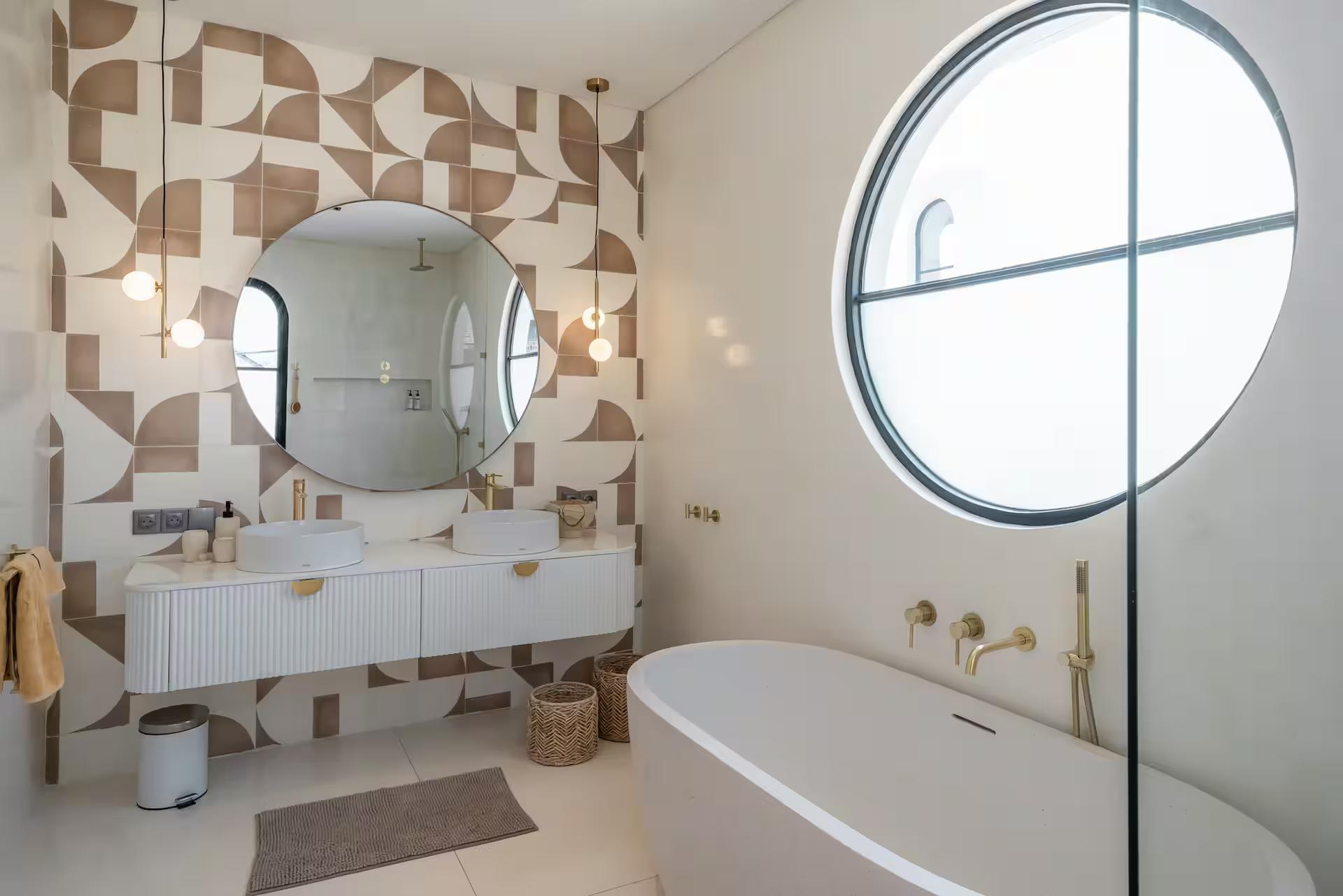 Freestanding tub below a round porthole window in the tan-and-white tiled master bathroom