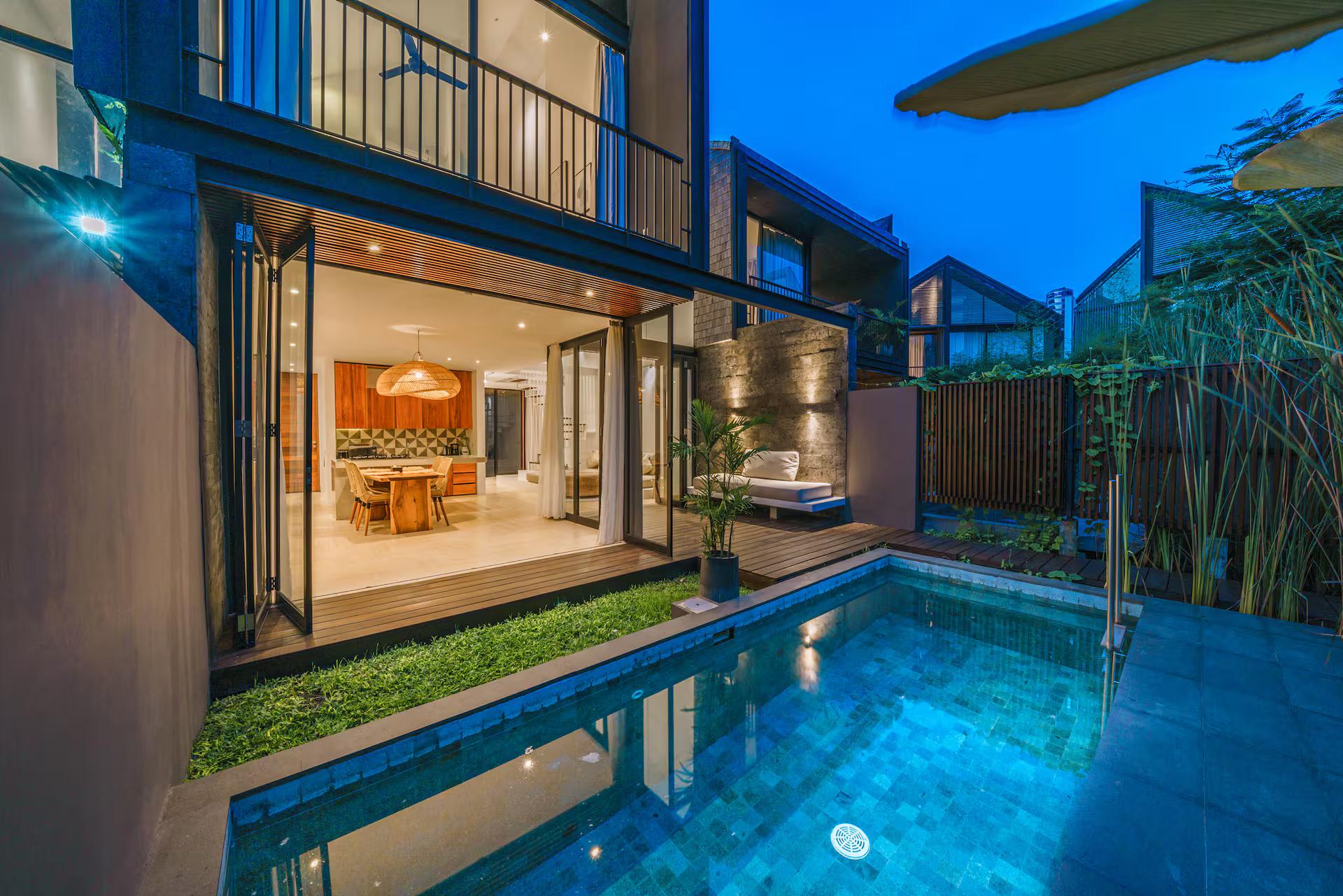 Villa RIZR at dusk, the lit pool reflecting the open kitchen and the upper-floor balcony