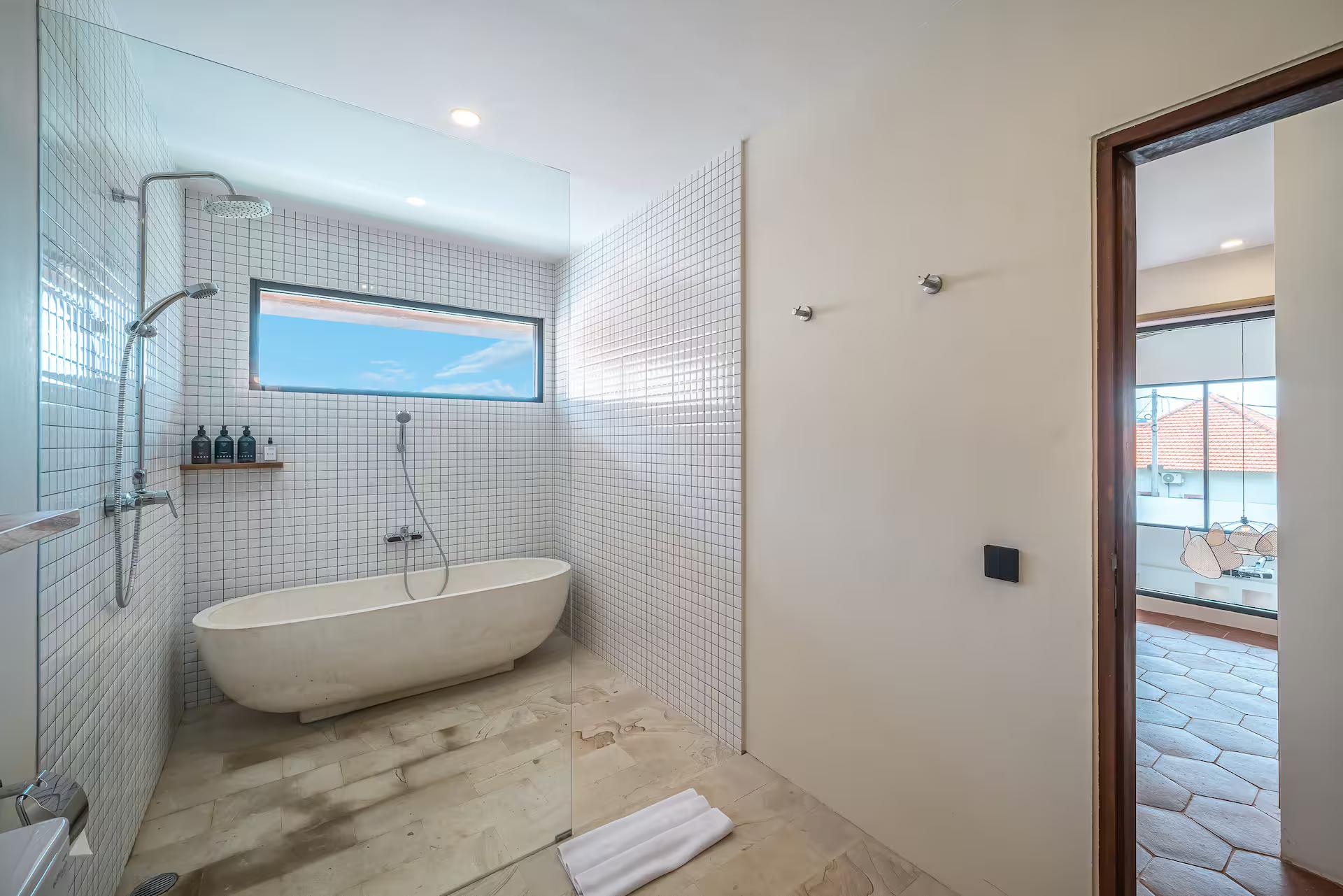 Freestanding stone soaking tub in a white mosaic-tiled wet room with a slot window