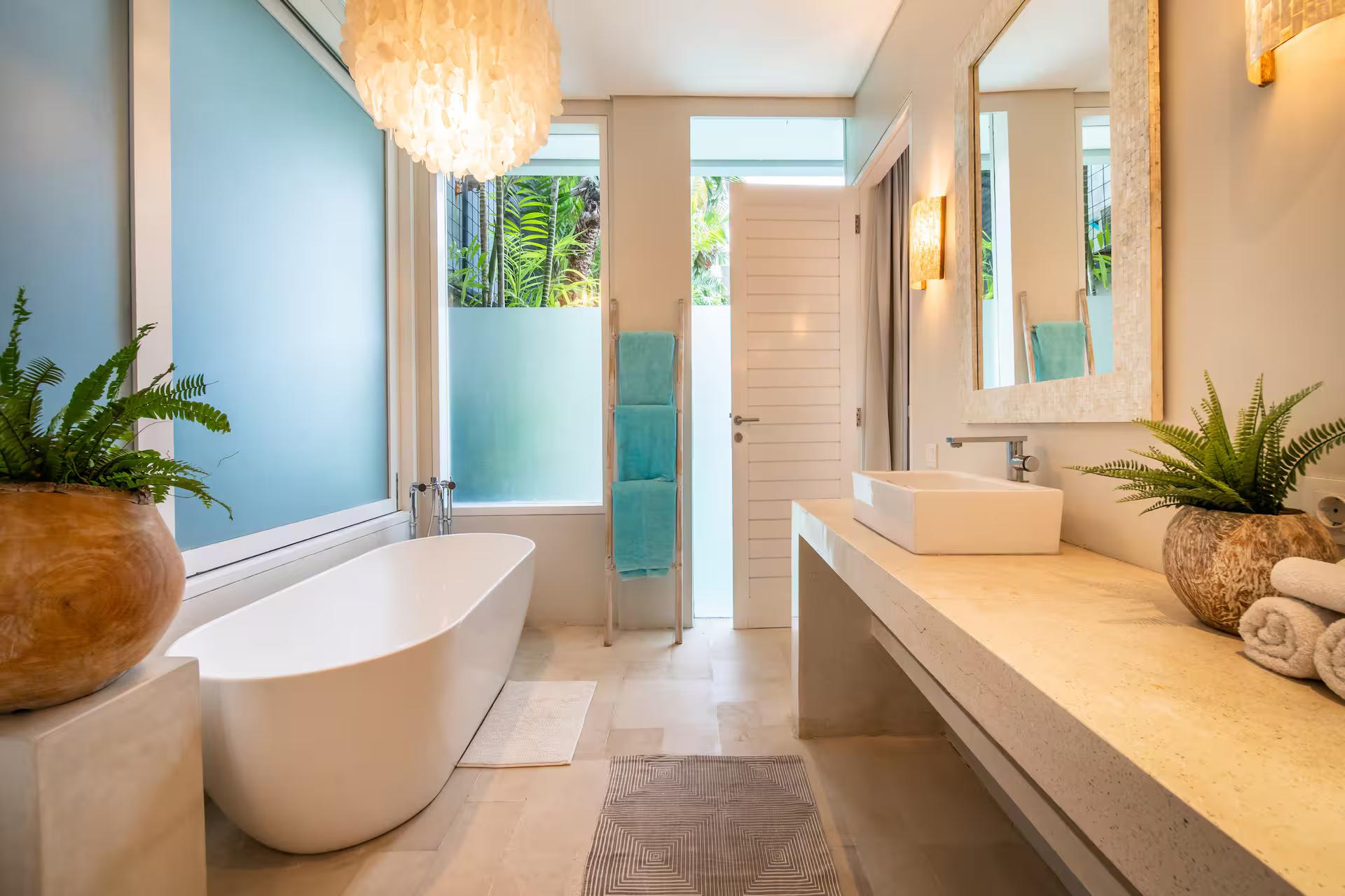 Freestanding oval tub against a teal wall with a long travertine vanity and potted ferns
