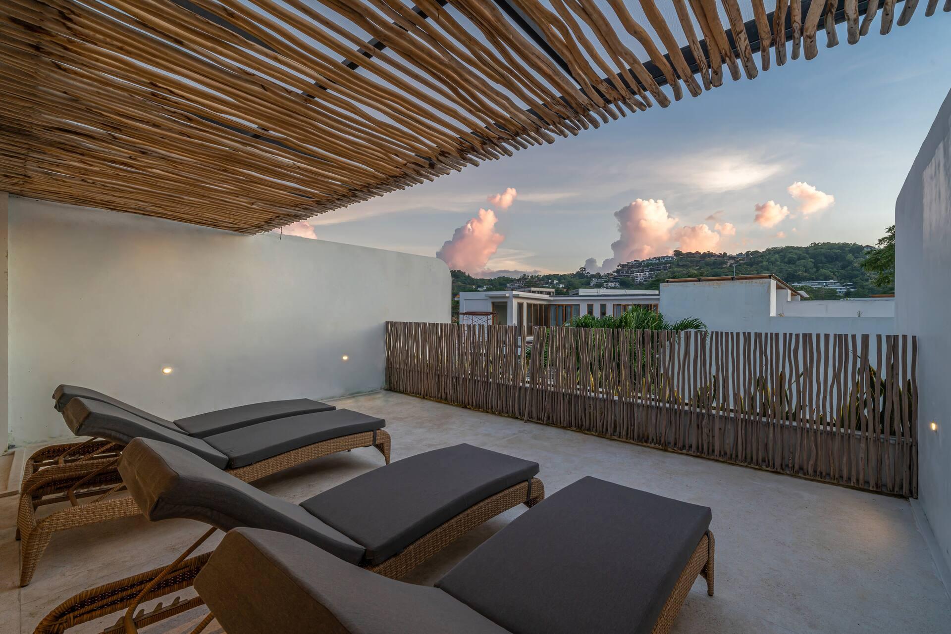 Roof terrace at sunset with grey sun loungers under a reed pergola and hills on the horizon