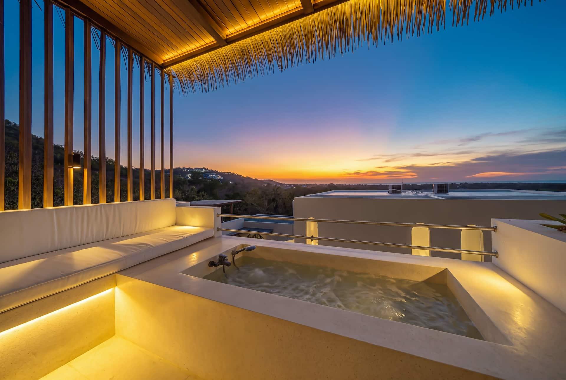 Built-in outdoor bathtub on the balcony at sunset, under a thatched canopy with hills beyond