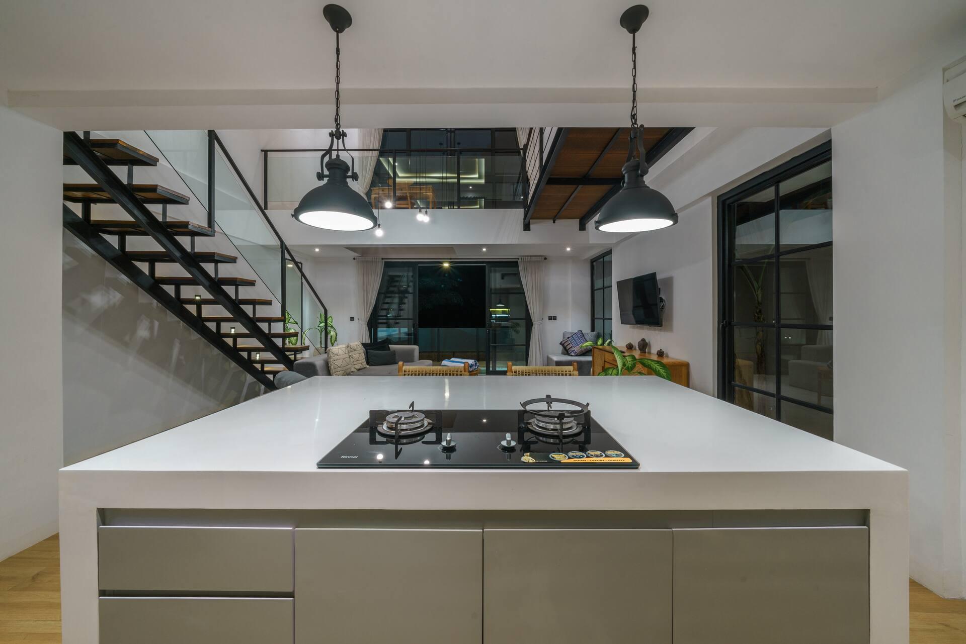 White kitchen island with a gas hob beneath black pendant lights, with the staircase behind