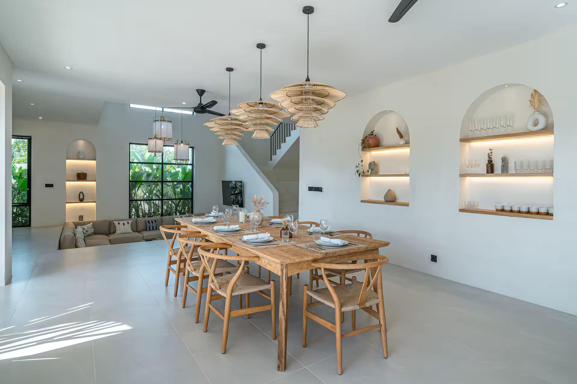 Long timber dining table under woven pendants, lit arched niches down the limewashed wall
