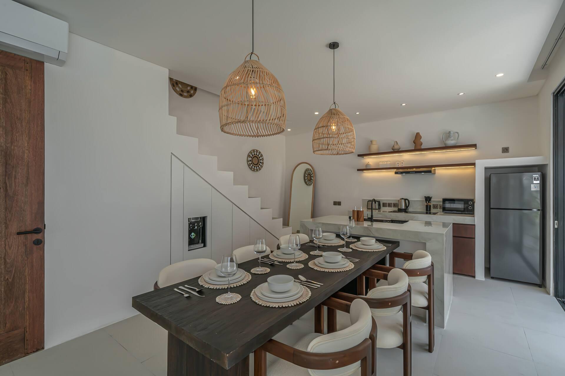 Dark timber dining table laid for six under three rattan pendants, the stone island behind