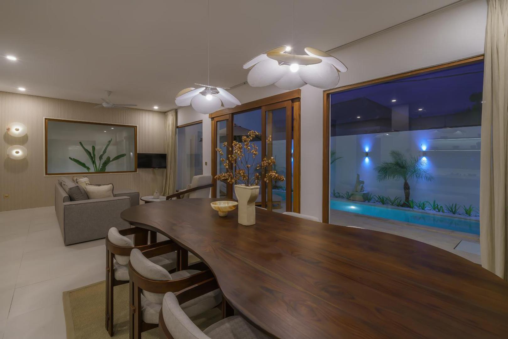 Curved live-edge dining table under petal pendants, the lit pool beyond the glass