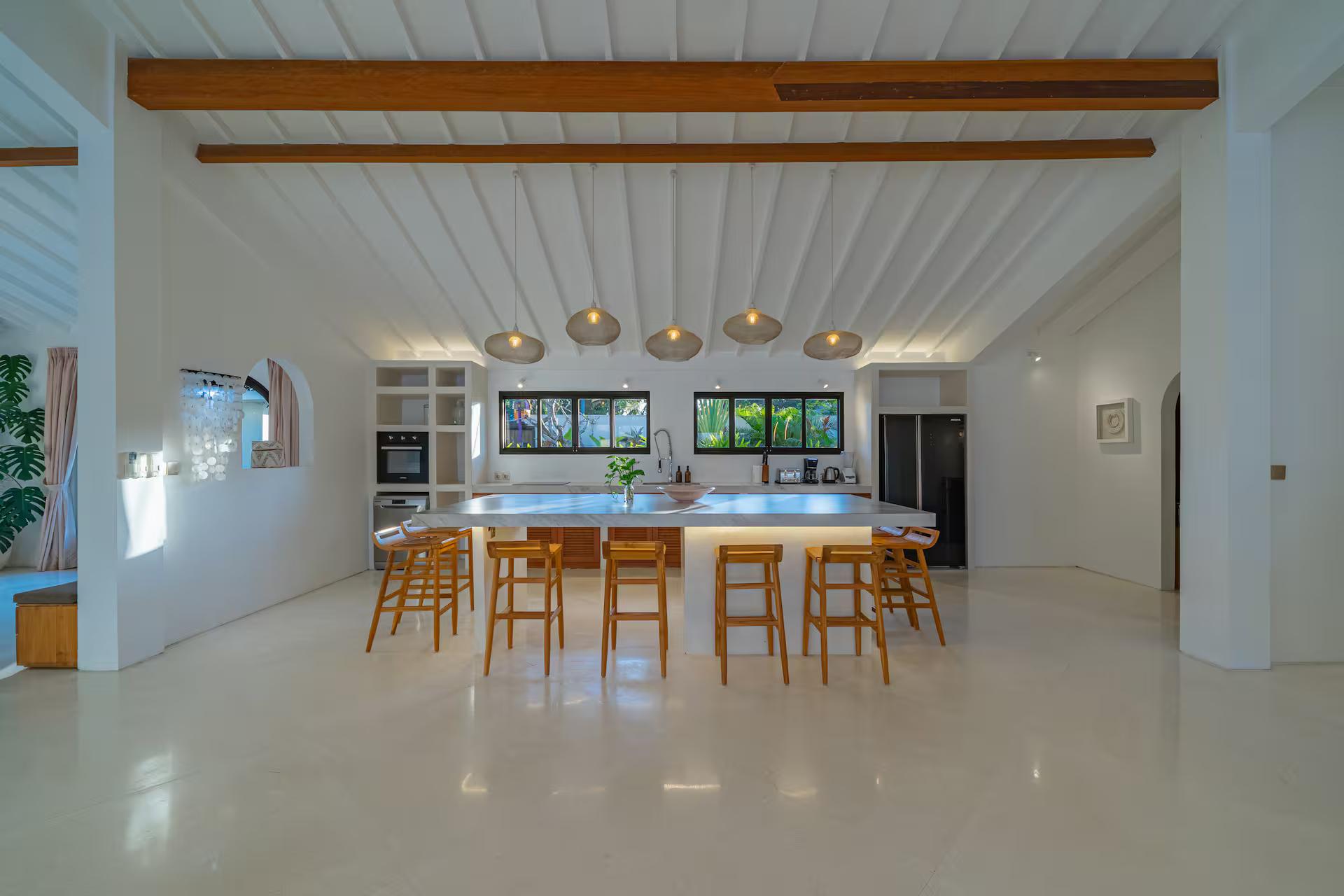 Long white kitchen island with pale timber bar stools under a line of brass dome pendants