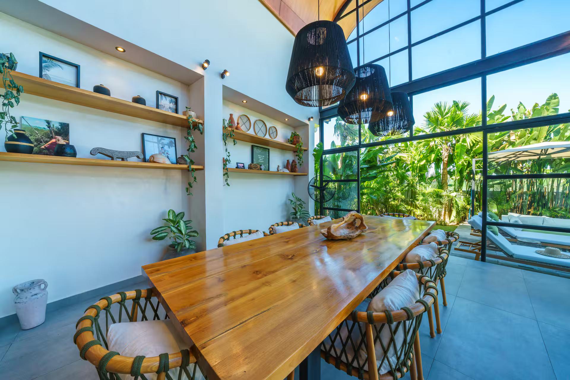 Long timber dining table with cane-backed chairs beside the black-framed glass wall