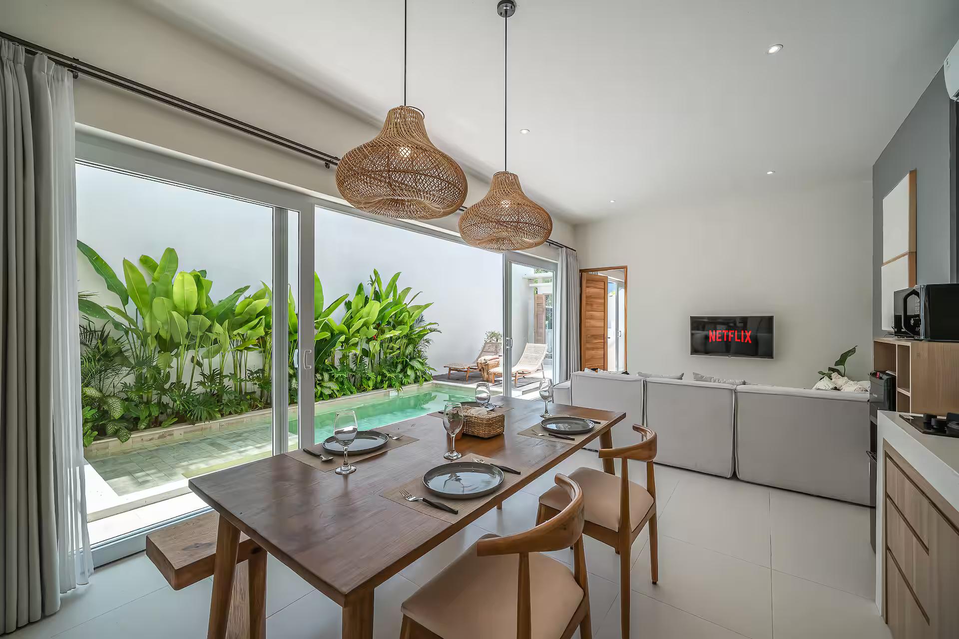 Timber dining table for four under rattan pendants, the pool bright through the glass doors